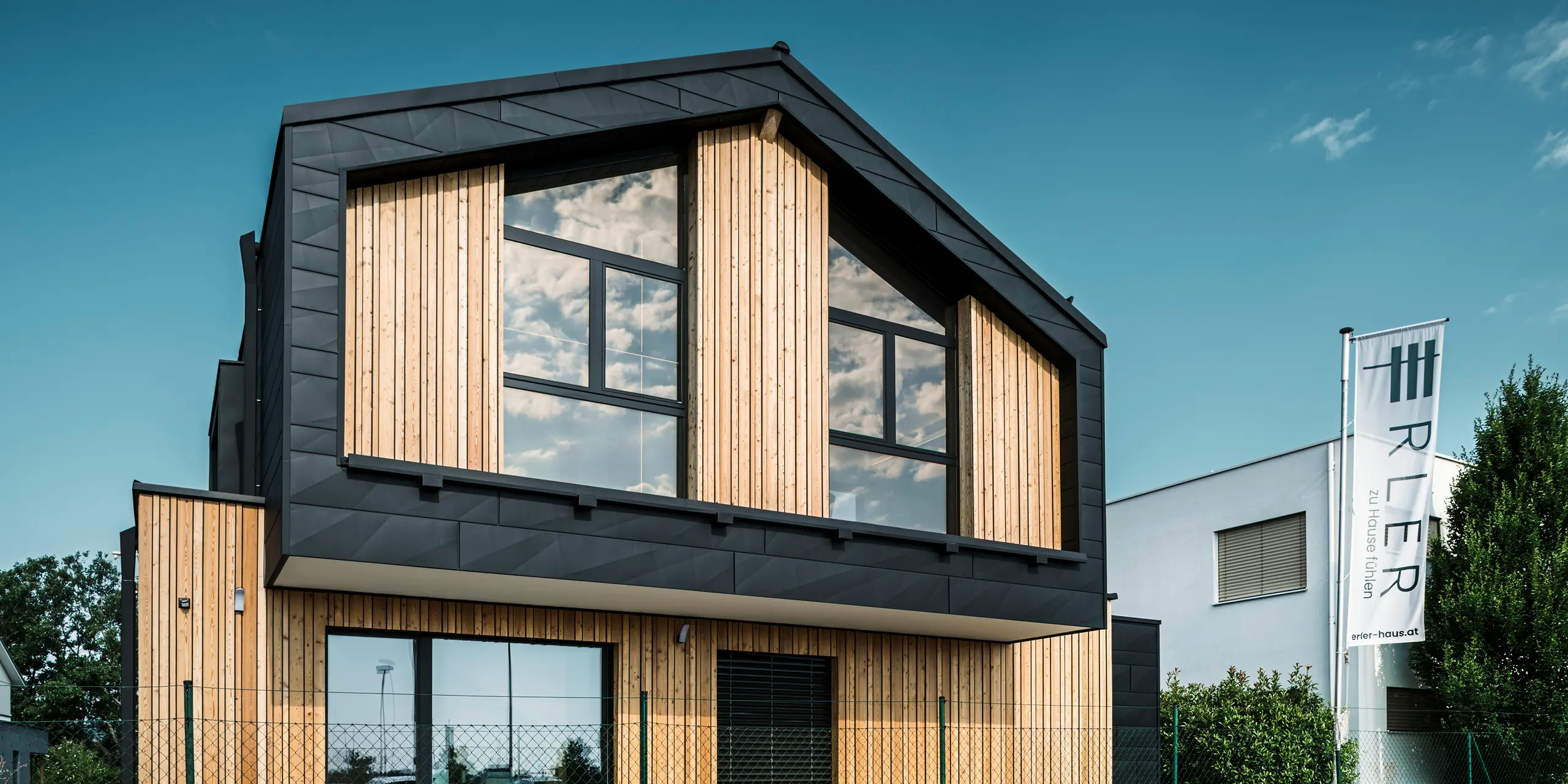 Čelna stran vzorčne hiše Autark v centru montažnih hiš Blaue Lagune v Vösendorfu (Avstrija). Fasada združuje navpične lesene elemente s črnimi aluminijastimi paneli PREFA Siding.X. Velike steklene površine na čelni strani odsevajo nebo in hiši dajejo sodoben, s svetlobo prežet videz. Zasnova poudarja harmonijo med lesom, steklom in aluminijem ter izraža sodobno in trajnostno arhitekturo.