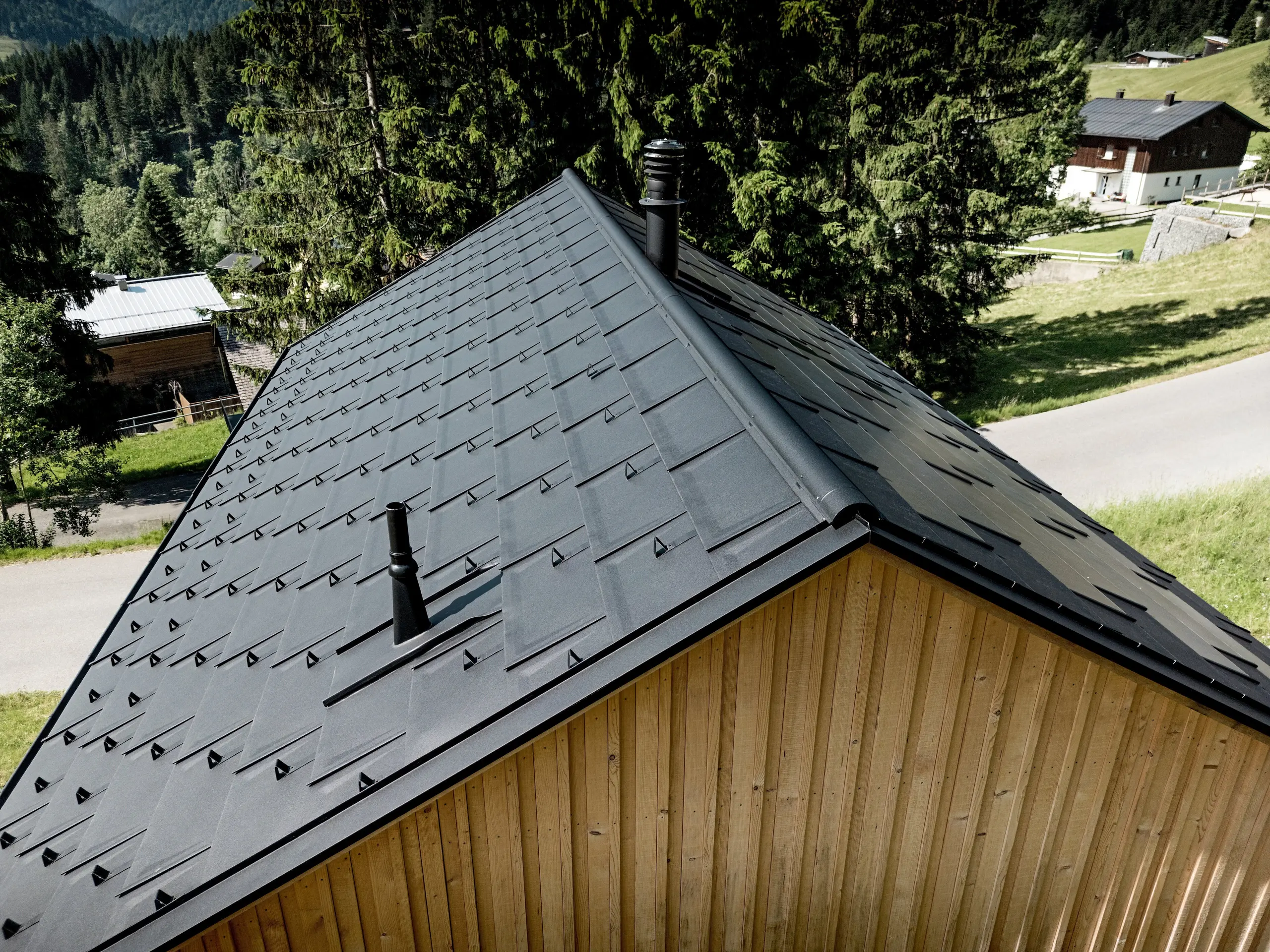 Dinamični bližnji posnetek prikazuje značilno strešno površino hiše Immi pred zelenimi gorskimi travniki in temnimi smrekami. PREFA strešne plošče R.16 v barvi P.10 črna, natančne detajlne rešitve in snežni lovilci tvorijo močno, enotno podobo strehe. Lahka, robustna aluminijasta strešna kritina je odporna proti alpskim vremenskim razmeram ter velikim obremenitvam s snegom ter leseno hišo v Ebnitu trajno ščiti in zahteva le malo vzdrževanja.