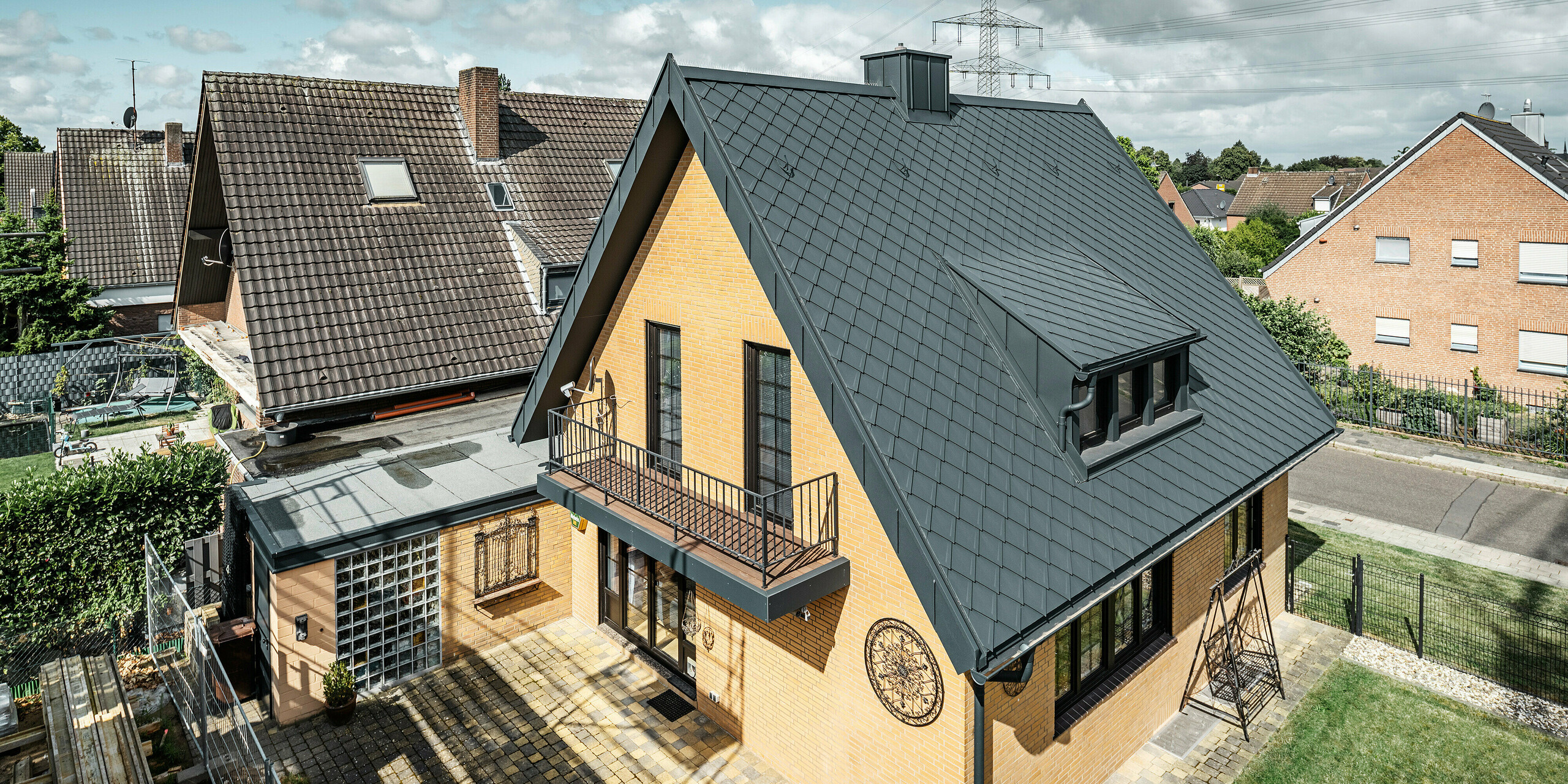 Enodružinska hiša v Tönisvorstu s koničasto dvokapno streho, prekrita s PREFA strešnimi rombi 29×29 v P.10 antracitni barvi. Streha ima jasne linije in strukturo. Žleb in odtočna cev sta barvno usklajena in se brezhibno prilegata dizajnu. Za hišo so značilne rumene opečne stene, ki so v kontrastu s temnimi strešnimi elementi, zunanjo podobo pa dopolnjujeta balkon in manjše gospodarsko poslopje.