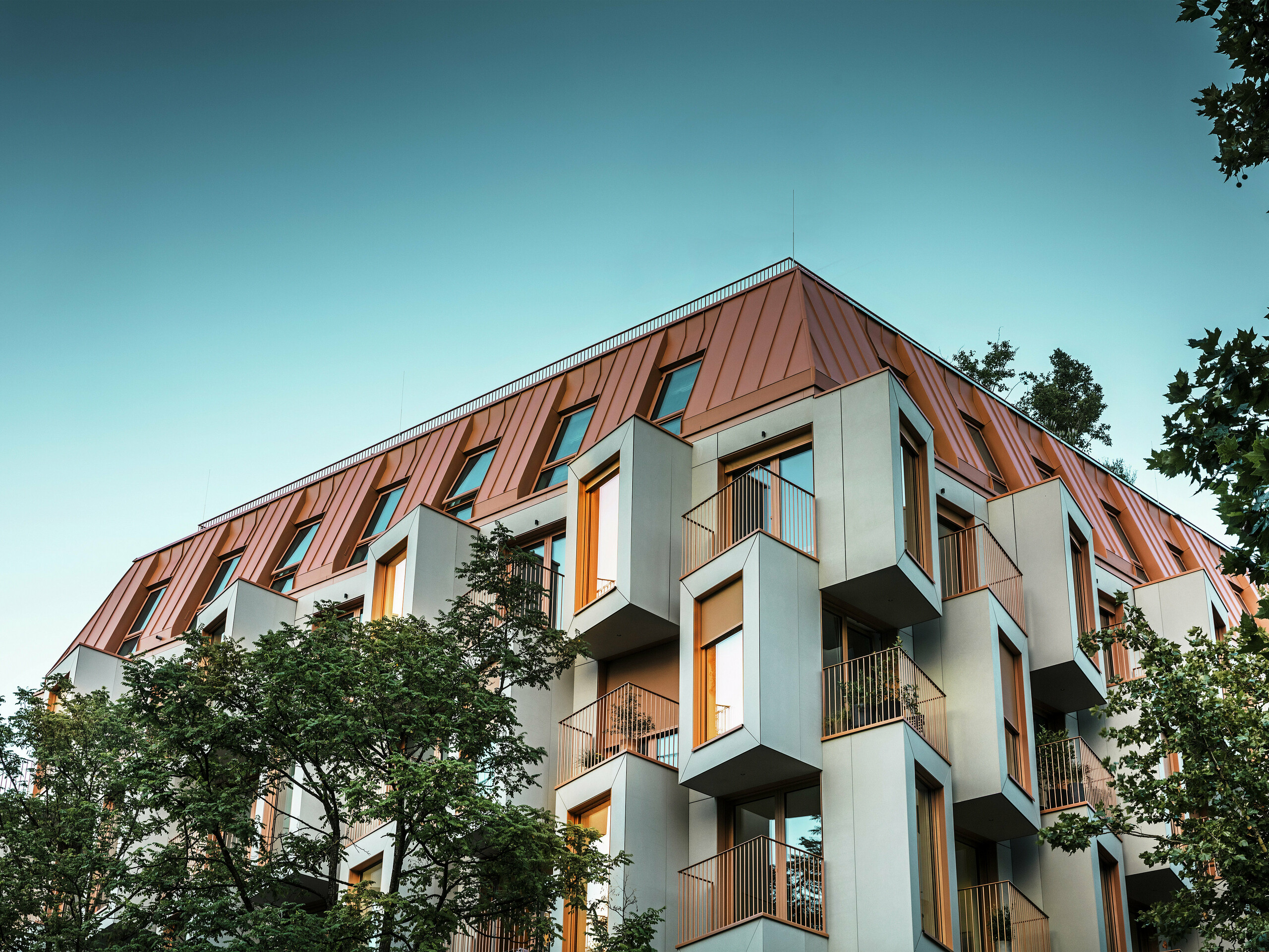 Arhitekturno izjemna fasada stanovanjskega projekta Van B v Münchnu s presenetljivimi erkerji in aluminijasto streho PREFALZ v posebni barvi IGP Orange HDP. Ob mraku se ob pogledu od spodaj pokažejo luči v oknih. Sodobna strešna rešitev združuje največjo odpornost proti vremenskim vplivom z elegantnim videzom in postavlja barvne poudarke v mestni prostor. Preplet betona iz steklenih vlaken, bakreno-oranžnih okenskih okvirjev in aluminijaste strehe ustvarja dinamičen, trpežen in trajnosten stavbni ovoj - idealen za gradnjo, usmerjeno v prihodnost.