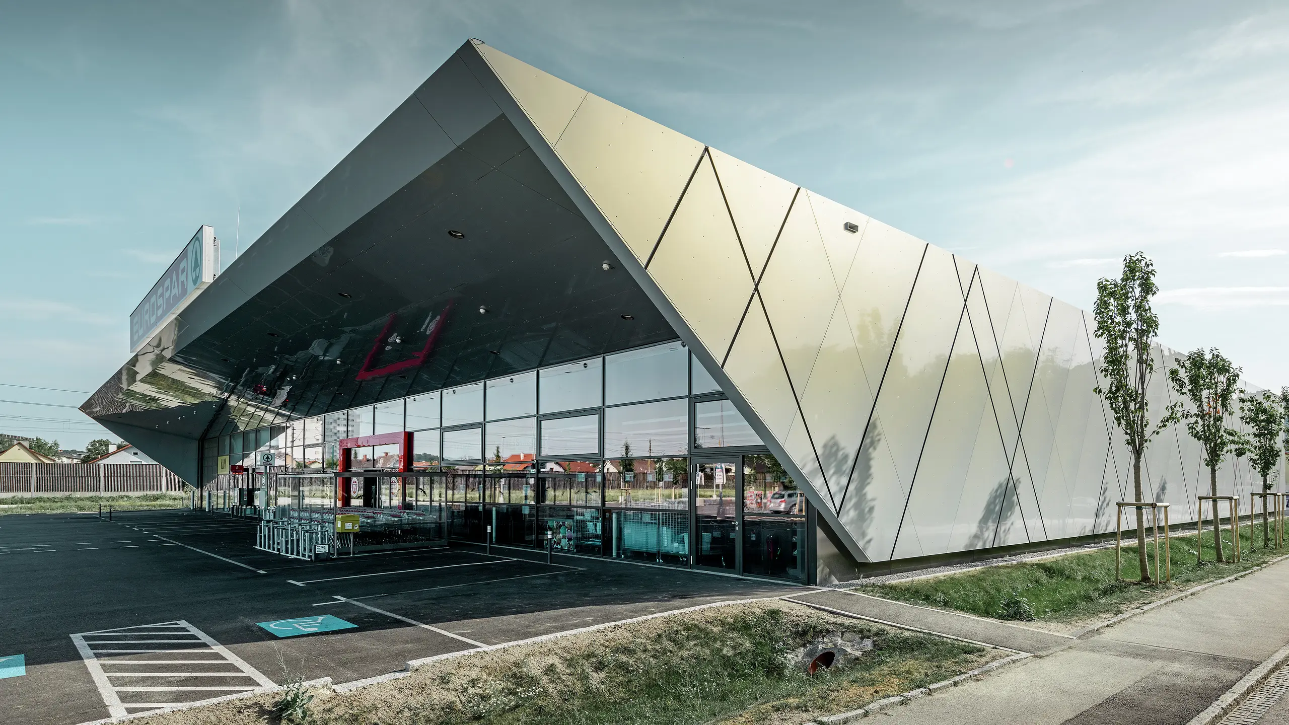 Vhodni prostor supermarketa EUROSPAR združuje velike steklene površine z izrazito PREFA aluminijasto fasado. Sijajna površina posebne barve Silver Gold poudarja visokokakovostno, v prihodnost usmerjeno arhitekturo supermarketa. Aluminijaste kompozitne plošče se v soncu bleščijo v različnih barvah, odvisno od kota pogleda.