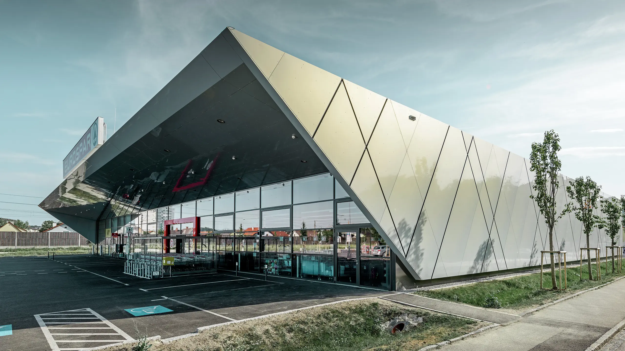 Vhodni prostor supermarketa EUROSPAR združuje velike steklene površine z izrazito PREFA aluminijasto fasado. Sijajna površina posebne barve Silver Gold poudarja visokokakovostno, v prihodnost usmerjeno arhitekturo supermarketa. Aluminijaste kompozitne plošče se v soncu bleščijo v različnih barvah, odvisno od kota pogleda.