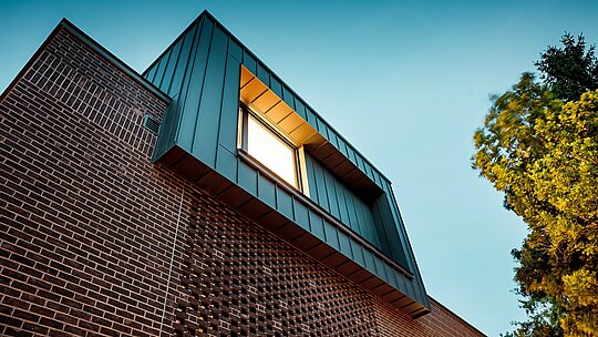 Fotografija prikazuje sodobno oblikovanje fasade večgeneracijske vile v Budimpešti iz dinamičnega pogleda od spodaj. Izstopajoči kubus iz PREFALZ aluminija v barvi P.10 antracit tvori izrazit kontrast s temno rdečimi opekami pod njim. Natančna izvedba navpičnih kotnih stoječih zgibov poudarja jasne linije in izpostavlja visoko kakovost PREFA aluminijastih izdelkov. Topla svetloba ustvarja močno prepletanje kovine, klinkerja in senc, ki impresivno poudarja inovativen, brezčasen značaj stavbe.