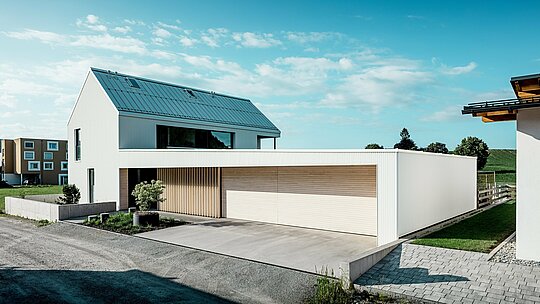 Slika prikazuje moderno enodružinsko hišo v Freistadtu, ki se odlikuje po preprosti, geometrijski obliki in kombinaciji bele PREFALZ aluminijaste fasade in lesene obloge. Lesena obloga na garažnih vratih ponuja topel, naraven kontrast in se popolnoma vklaplja v moderno oblikovanje. Streha, prav tako pokrita s PREFALZ, zagotavlja neprekinjeno, vremensko odporno površino. Ta kombinacija aluminija, ki je enostaven za vzdrževanje, in lesa ustvarja estetsko privlačno in funkcionalno rešitev za trajnostno arhitekturo. Prostran zeleni zunanji prostor in jasno nebo v ozadju poudarjata moderno kakovost življenja, ki jo ponuja ta hiša.