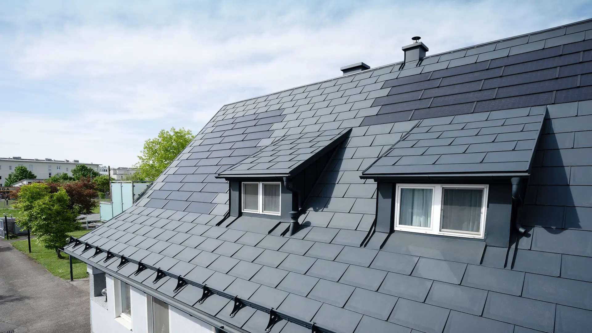 Približek strehe, pokrite z velikimi solarnimi strešnimi ploščami PREFA in strešno ploščo PREFA R.16 v barvi P.10 antracitna. Vidni sta dve strešni okni. Solarne strešne plošče so enakomerno razporejene po strehi in zagotavljajo moderno, trajnostno rešitev.