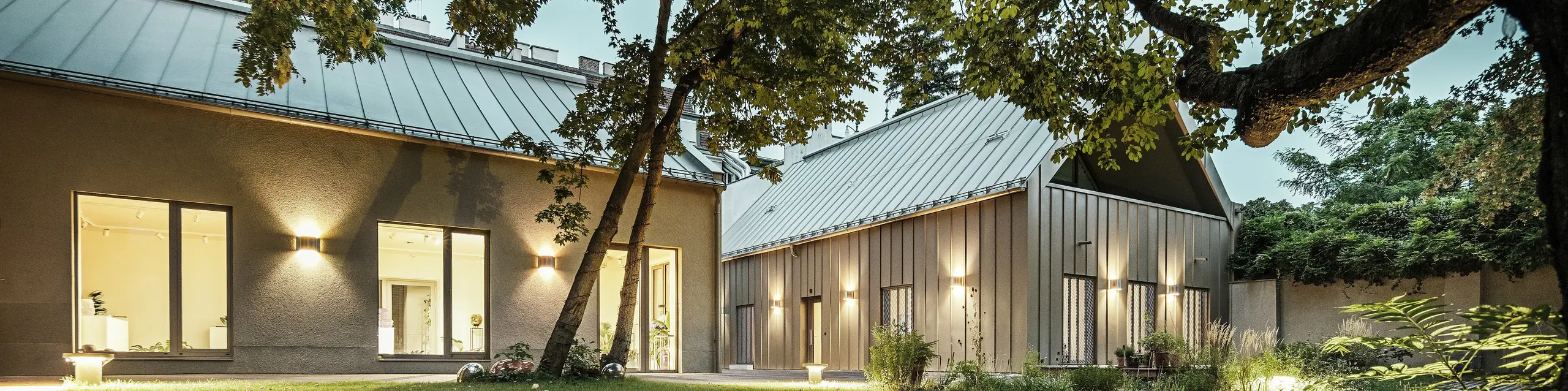 Večerni pogled na Studio Comploj na Dunaju, obdan z drevesi in zelenimi površinami. Fasada in streha iz aluminija PREFALZ v odtenku P.10 Prefa bronasta se harmonično vključujeta v okolico.