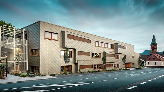 Posnetek z ravni ceste prikazuje nov vrtec v Alzenau v zanimivem dialogu z zgodovinsko mestno kuliso, med katero v ozadju izstopa značilna cerkev. Sodobna fasada iz bronastih PREFA fasadnih rombov 44 × 44 daje stavbi eleganten, skoraj skulpturen videz. Natančno nameščena okna, navpične lesene lamele in zamaknjene fasadne površine ustvarjajo globino in ritem. Na levi je vidno filigransko stopnišče z belim kovinskim ogrodjem, ki daje vtis sodobne lahkotnosti. Jasna oblika, trajnostna izbira materialov in visokokakovostna aluminijasta fasada stavbo spreminjajo v vzorčni projekt za urbanistično integracijo in prihodnostno usmerjeno gradnjo s PREFA sistemi.