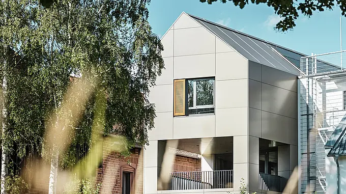 Slika prikazuje moderno mestno hišo v Alzenauu, katere fasada je izdelana iz aluminijastih kompozitnih plošč PREFA v elegantnem bronastem odtenku. Ostrorobi del strehe in velika okna poudarjajo sodobni značaj stavbe. V ospredju stavbo obdajajo drevesa in zelenice, mehka osvetlitev in jasno nebo pa poudarjata sodobni urbani prostor.