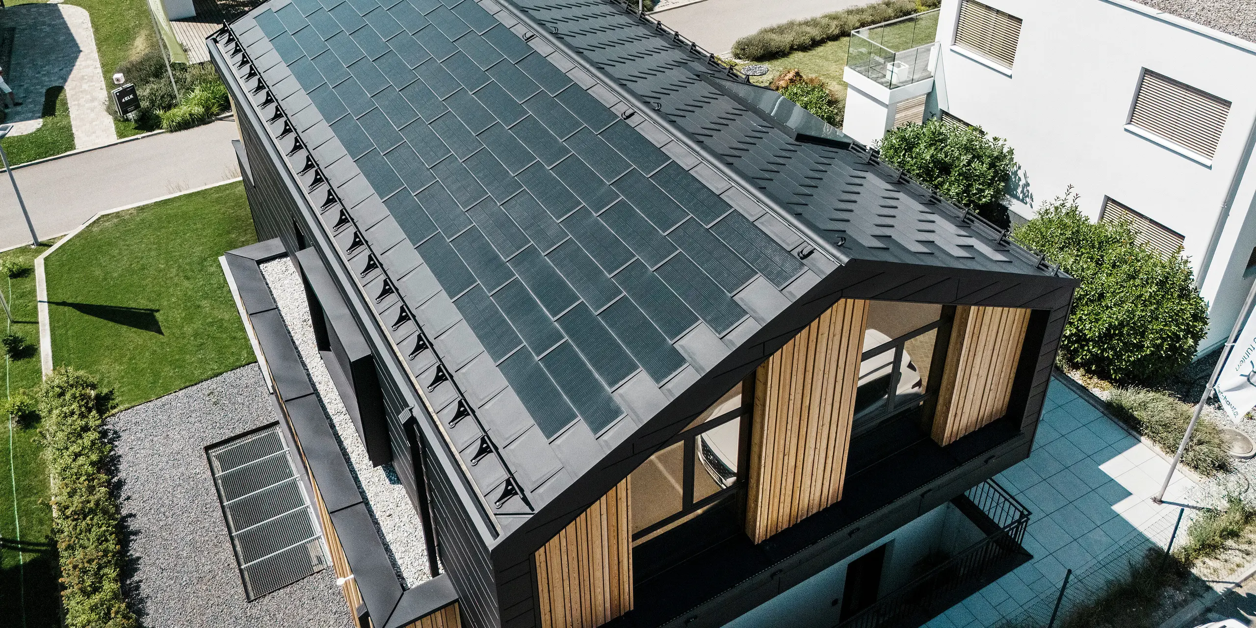 Pogled iz zraka na vzorčno hišo Autark v centru montažnih hiš Blaue Lagune v Vösendorfu (Avstrija) z robustno aluminijasto streho PREFA. Streha je prekrita s solarnimi strešnimi ploščami PREFA in strešnimi ploščami R.16 v barvi P.10 črna. Vgrajena fotonapetostna naprava omogoča trajnostno pridobivanje energije s pomočjo sončne svetlobe. Lesena obloga na čelni strani dopolnjuje sodoben videz hiše.