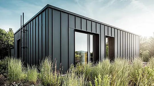 Die Aufnahme zeigt die moderne, kubische Dachterrasse der Budapester Mehrgenerationen-Villa, verkleidet mit hochwertigem PREFALZ Aluminium in P.10 Anthrazit. Die klar strukturierten Winkelstehfalze erzeugen eine elegante, vertikale Linienführung, die durch das weiche Sonnenlicht besonders betont wird. Umgeben von dichter, natürlicher Bepflanzung entsteht ein spannender Kontrast zwischen moderner Metallarchitektur und üppigem Grün. Die langlebigen, witterungsbeständigen PREFA Aluminiumprodukte unterstreichen die hochwertige Ausführung und machen die Dachterrasse zu einem markanten, architektonischen Highlight.