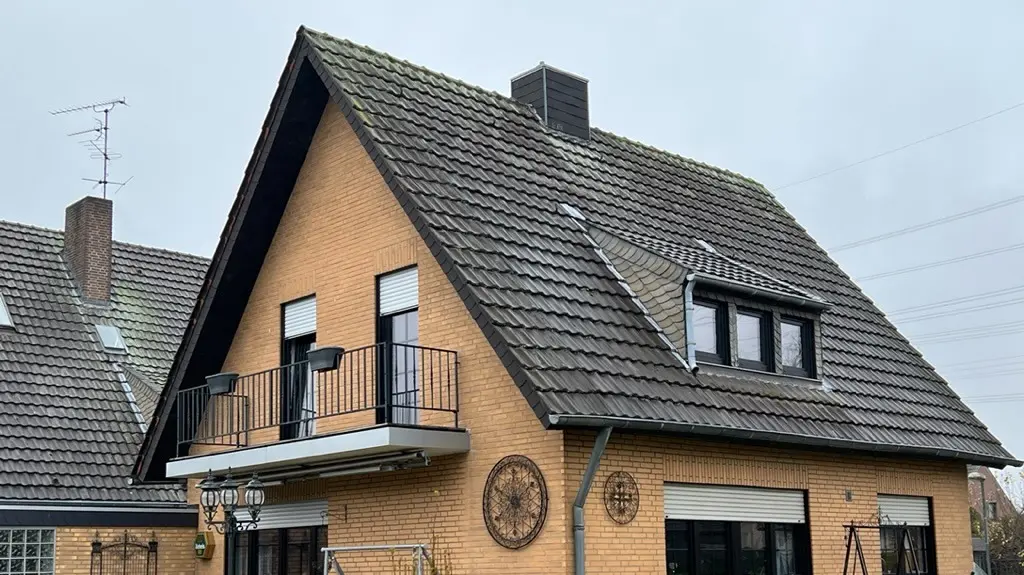 Enodružinska hiša iz rumene opeke s strmim dvokapnim strešnim pokritjem in temno strešno kritino pred obnovo strehe; na strani vrta z balkonom, strešno lopo, teraso s sedežno garnituro in travnikom v ospredju.