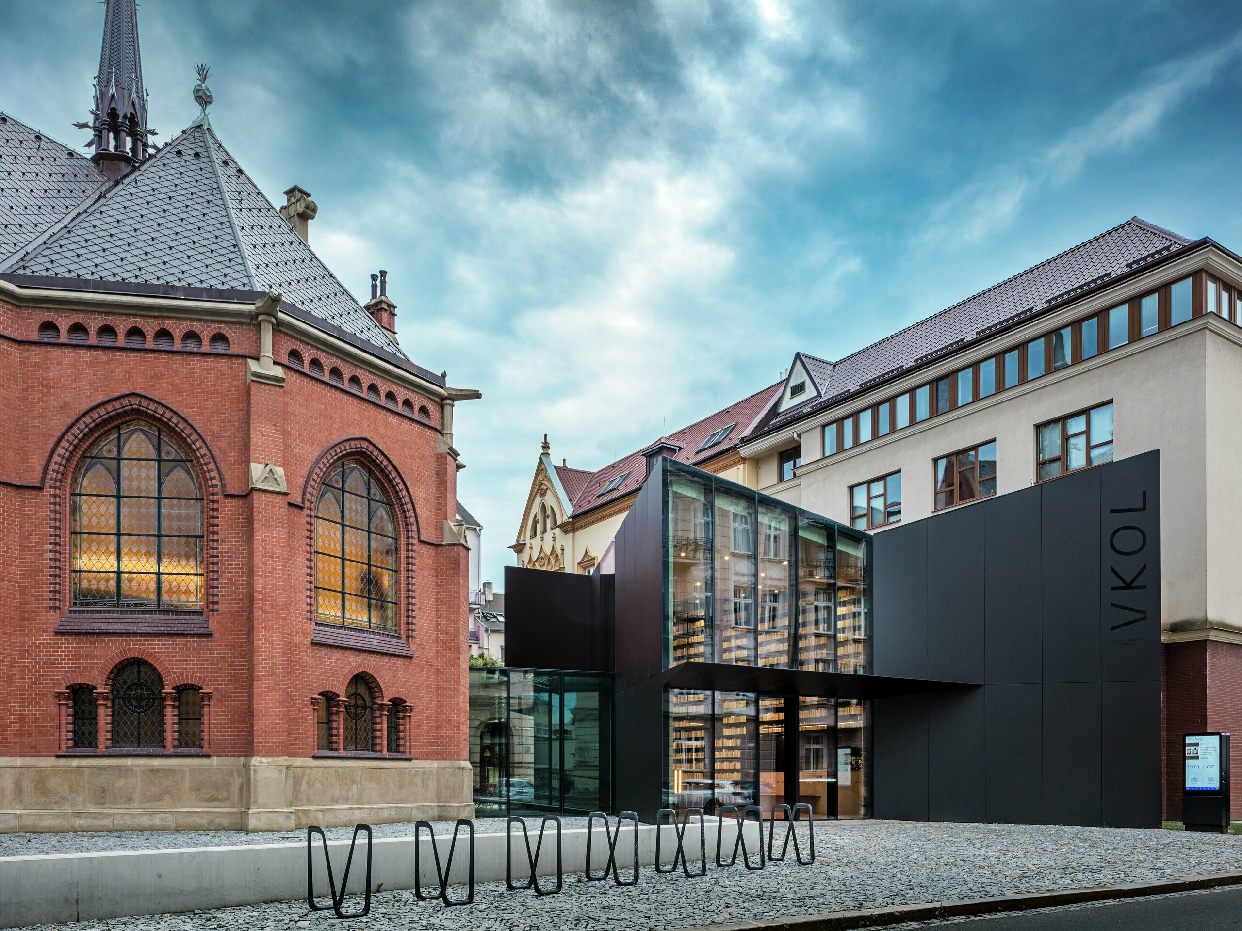 Slika prikazuje fascinantno povezavo zgodovinske in sodobne arhitekture v Olomucu. Na levi strani stoji veličastna Rdeča cerkev s svojimi značilnimi gotskimi okni in rdečimi opečnimi zidovi, medtem ko nova stavba na desni strani tvori kontrast s svojo moderno črno fasado iz aluminijastih kompozitnih plošč PREFABOND. Steklena fasada nove stavbe omogoča vpogled v inovativno zasnovo knjižnice in daje kompleksu eleganten, odprt značaj. Natančna obdelava plošč PREFABOND zagotavlja enoten videz, ki se harmonično vklaplja v zgodovinsko okolico in hkrati daje sodoben pečat. Arhitektura tega projekta popolnoma združuje tradicijo in inovativnost, pri čemer materiali ne prepričajo le estetsko, ampak zagotavljajo tudi dolgo življenjsko dobo in trajnost.