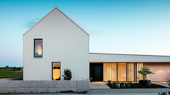 Das Bild zeigt das beeindruckende Einfamilienhaus in Freistadt, das sich durch seine markante, geometrische Architektur auszeichnet. Die Fassade des Gebäudes besteht aus präzise gefaltetem, reinweißem PREFALZ, welches dem Haus seine moderne Eleganz verleiht. Die vertikale Struktur der Fassade und das klare, minimalistische Design unterstreichen die hochwertige Materialwahl und sorgen für eine langlebige, witterungsbeständige Oberfläche. Das steile Satteldach, das ebenfalls mit PREFALZ verkleidet ist, schützt vor den Elementen und sorgt gleichzeitig für eine harmonische Gesamtästhetik. Durch die klare Formensprache und die Verwendung von Aluminium werden sowohl ästhetische als auch funktionale Anforderungen optimal erfüllt. Die lichtdurchfluteten Fenster und das offene Eingangsportal wirken einladend und setzen einen spannungsvollen Kontrast zur modernen Fassadengestaltung. Die Entscheidung für PREFALZ unterstreicht den architektonischen Anspruch und stellt eine nachhaltige, pflegeleichte Lösung für langlebige Bauprojekte dar.