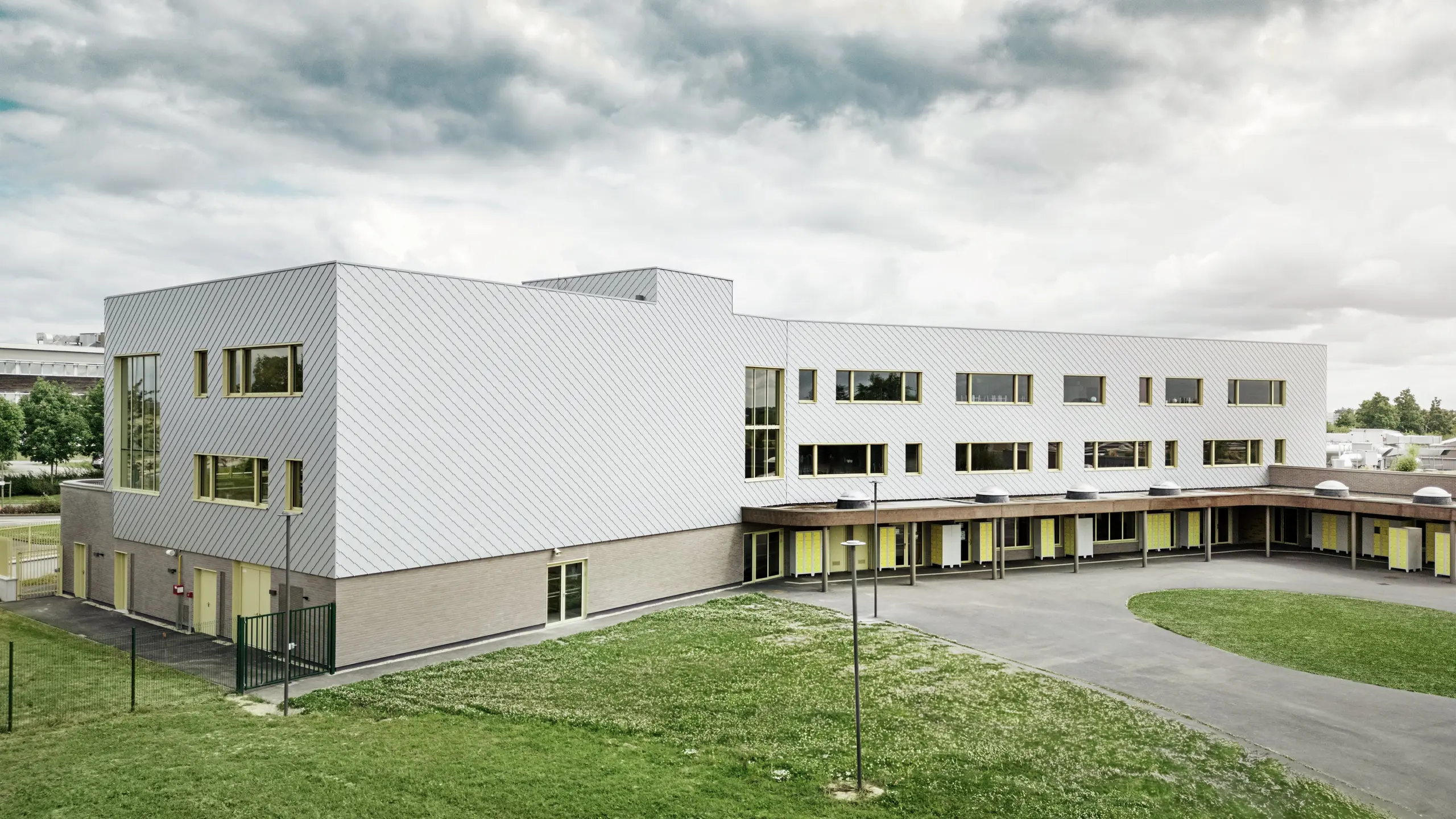 Slika prikazuje zadnjo stran šole Collège Léontine Dolivet. V ospredju je prostrano, asfaltirano šolsko dvorišče z jasnimi linijami in odprtimi površinami, ki se lahko uporabljajo za odmore in prostočasne dejavnosti. Nekaj zelenih površin popestri okolico. V ozadju se dviga moderna šolska stavba z izrazito aluminijasto fasado iz kovinsko srebrnih PREFA fasadnih rombov, ki odbijajo svetlobo in ustvarjajo dinamičen učinek. Več velikih oken zagotavlja svetlost v notranjosti. Slika je posneta pri dnevni svetlobi, ob rahlo oblačnem nebu, kar še posebej poudarja kovinsko površino.