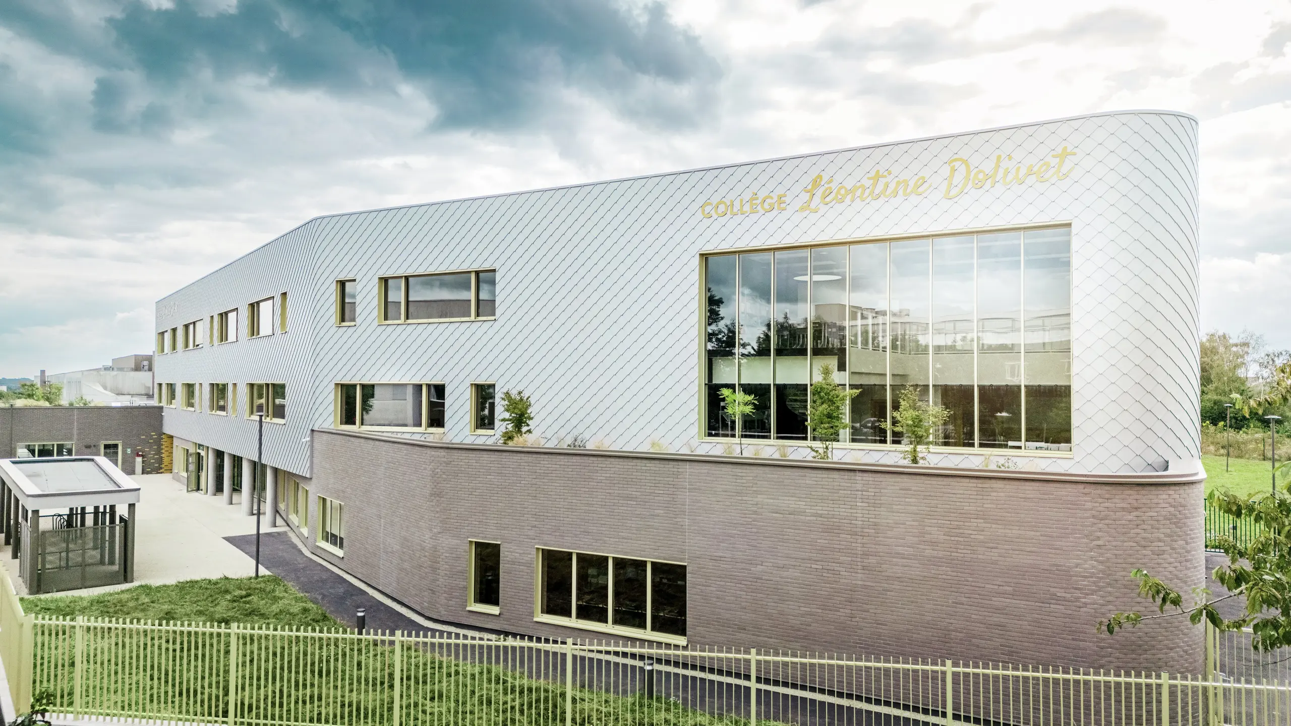 Stranski pogled na Collège Léontine Dolivet v Cesson-Sévigné z moderno aluminijasto fasado iz PREFA fasadnih rombov v kovinsko srebrni barvi. Diagonalna struktura ustvarja dinamičen videz, velike steklene površine in rumene polkna pa dodajajo zanimive poudarke. V ospredju so vidne zelene površine in ograja, ki obdajajo stavbo. Trajna fasadna rešitev PREFA združuje eleganco, odpornost proti vremenskim vplivom in trajnost – arhitekturni magnet za poglede z jasnimi linijami in spreminjajočim se kovinskim videzom.