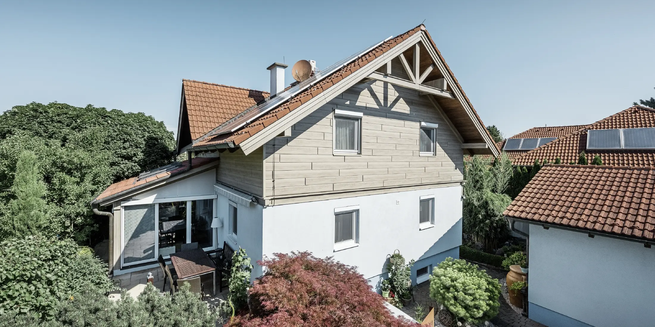 Enodružinska hiša s strmo streho in rdečo strešno kritino, kombinirana z moderno fasado. Zgornji čel je obložen z bež-sivimi aluminijastimi fasadnimi panelji PREFA, medtem ko ima pritličje belo fasado iz ometa. Na strehi so nameščeni solarni moduli. Hiša stoji v zelenem vrtu s teraso, vrtnim pohištvom in bujno vegetacijo, obkrožena z drugimi stanovanjskimi hišami s podobno strešno arhitekturo.