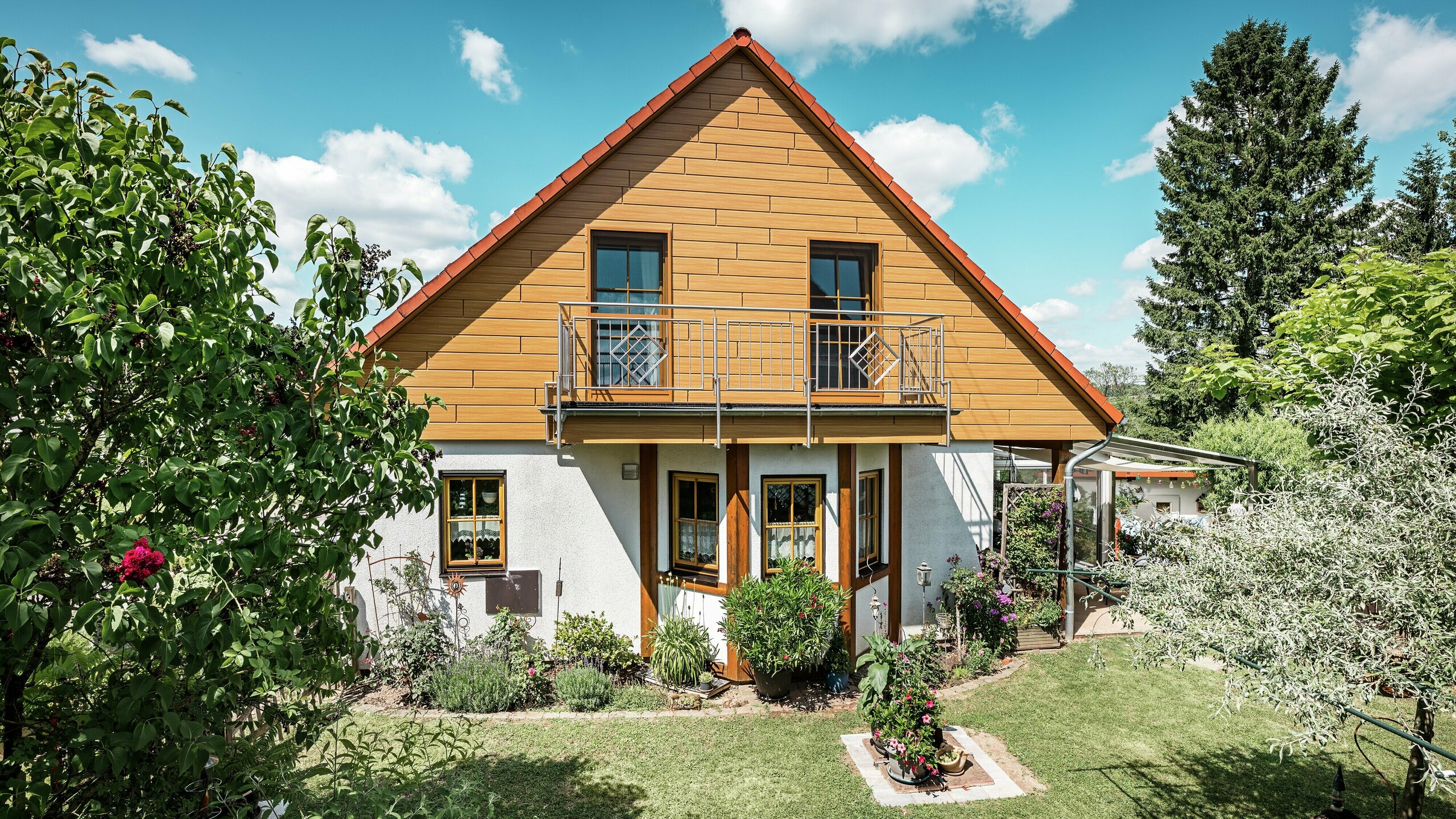 Na tej sliki obnoveni čelni del enodružinske hiše v Blieskastelu žari v topli barvi naravnega hrasta zahvaljujoč PREFA Sidingu. Visokokakovostna aluminijasta fasada z realističnim videzom lesa ustvarja močan vizualni poudarek in se impresivno ujema z rdečo streho iz strešnikov ter bujnim vrtom. Obkrožena s cvetjem, grmičevjem in bujno zeleno vegetacijo hiša deluje vabljivo in urejeno. PREFA površina združuje naraven videz s funkcionalnimi prednostmi, kot so visoka odpornost proti vremenskim vplivom, stabilnost in praktično brez vzdrževanja – estetski vrhunec s trajnostno dodano vrednostjo.