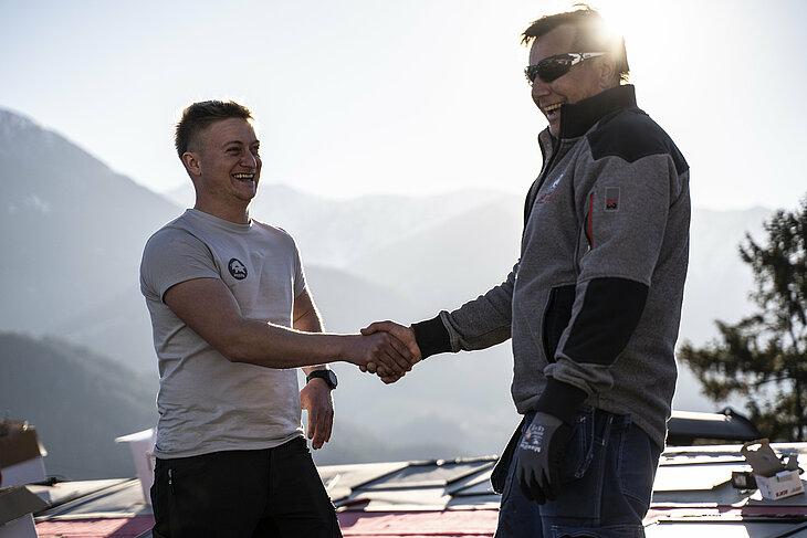 Na poševni strehi hiše stojita PREFA inštruktor in klepar, ki si segata v roke. Oba nosita profesionalna delovna oblačila in varnostno opremo. V ozadju je vidno nizko stoječe sonce in embalažne škatle PREFA aluminijastih izdelkov. Ta prizor simbolizira uspešno sodelovanje in strokovno izmenjavo z ročnim stiskom. PREFA inštruktor krovcu pomaga s svojim znanjem o obdelavi, tehniki in sistemih izdelkov. Slika predstavlja tesno sodelovanje podjetja PREFA z obrtniki – od usposabljanja do profesionalne izvedbe na gradbišču.