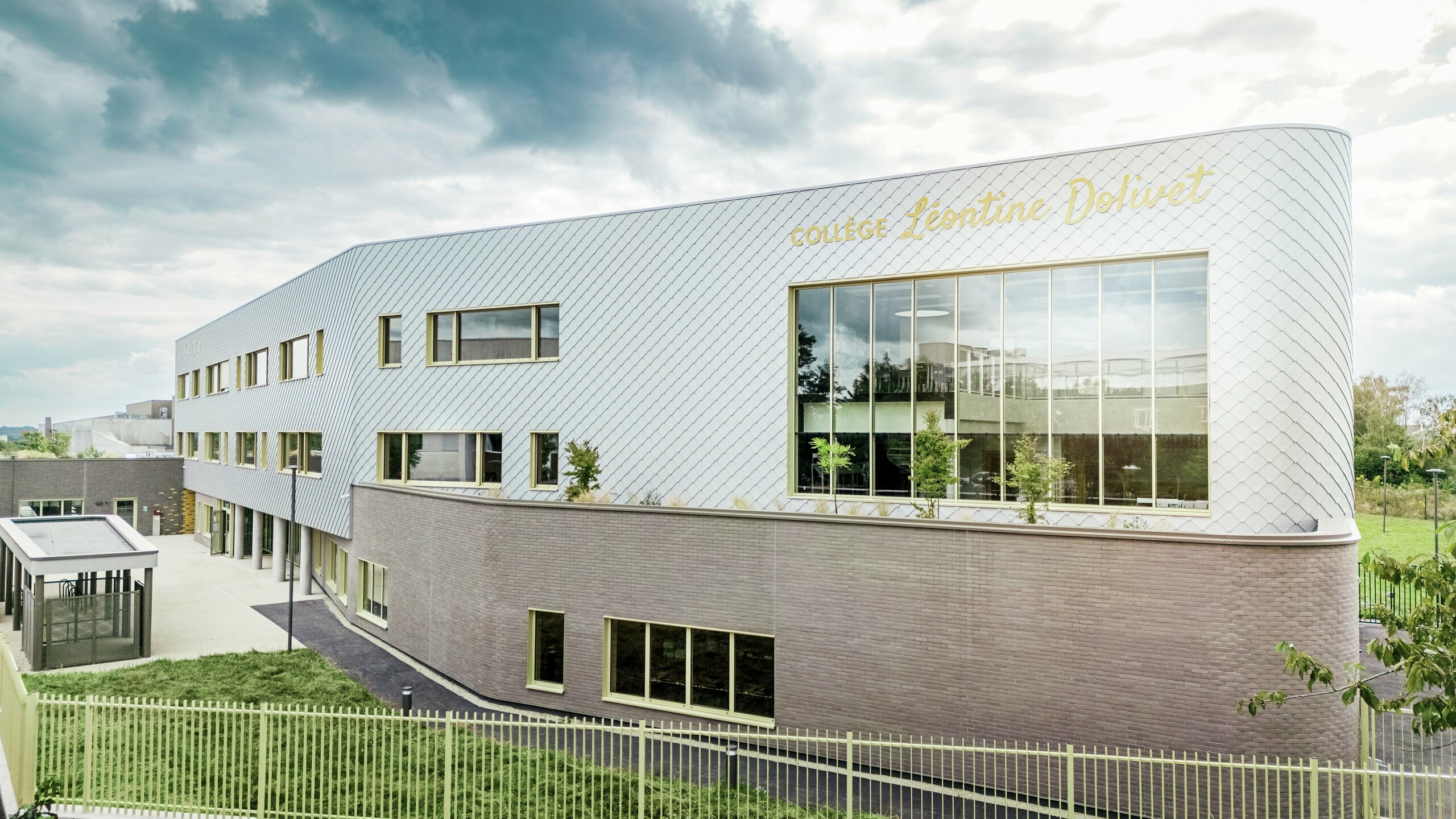 Stranski pogled na Collège Léontine Dolivet v Cesson-Sévigné z moderno aluminijasto fasado iz PREFA fasadnih rombov v kovinsko srebrni barvi. Diagonalna struktura ustvarja dinamičen videz, velike steklene površine in rumene polkna pa dodajajo zanimive poudarke. V ospredju so vidne zelene površine in ograja, ki obdajajo stavbo. Trajna fasadna rešitev PREFA združuje eleganco, odpornost proti vremenskim vplivom in trajnost – arhitekturni magnet za poglede z jasnimi linijami in spreminjajočim se kovinskim videzom.