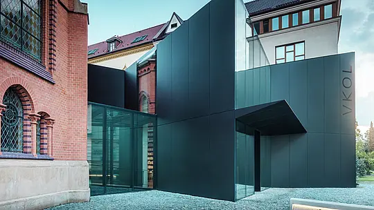 Slika prikazuje moderno razširitev knjižnice, ki se elegantno navezuje na zgodovinsko Rdečo cerkev v Olomouncu. Izrazita nova stavba navdušuje s fasado iz črnih PREFABOND aluminijastih kompozitnih plošč, ki je oblikovana v jasnih, geometrijskih oblikah in prepriča s precizno izdelanimi detajli. Steklene stene stavbe omogočajo prosojen pogled v notranjost, ki se harmonično vklaplja v zgodovinsko okolico. Sodelovanje sodobnih materialov in klasičnih arhitekturnih elementov ustvarja zanimivo vizualno povezavo med starim in novim. Inovativna uporaba plošč PREFABOND ne zagotavlja le sodobnega videza, ampak tudi dolgo življenjsko dobo in odpornost, ki stavbo ščitita za prihodnost.