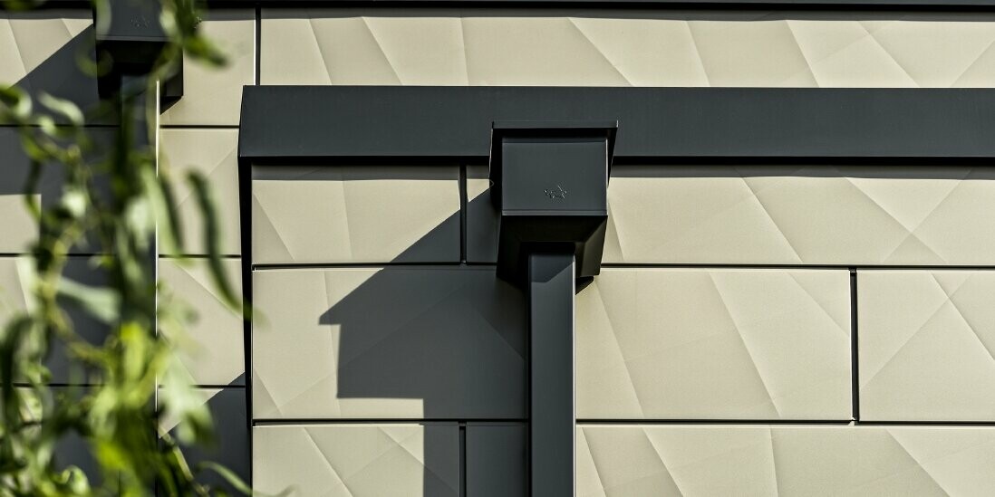 Moderna enodružinska hiša z ravno streho in ozkimi okni. Fasada je izdelana iz panelov PREFA Siding.X z nagubanim videzom v bronasti barvi. Priključki so bili izdelani v antracitni barvi. Za odvodnjavanje strehe je uporabljena pravokotna odtočna cev PREFA z ustreznim zbiralnim kotličkom v antracitni barvi.