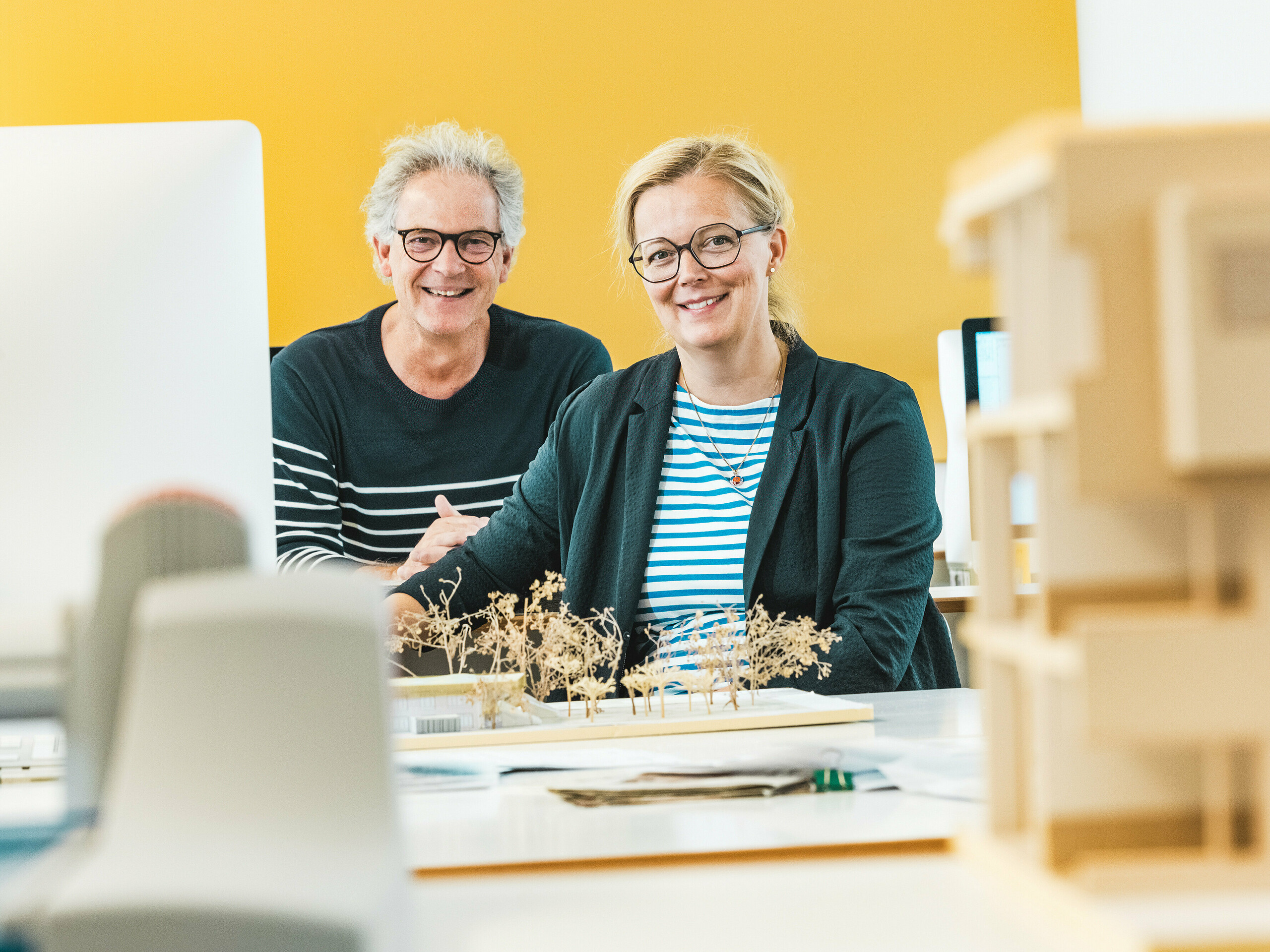 Arhitekta Claudia in Theo Härtner sedita vesela pred arhitekturnim modelom. Arhitekturna dvojica je realizirala vrtec v Alzenauu, futuristično hibridno stavbo s fasado iz PREFA aluminija. V ospredju arhitekturnega modela so miniaturna drevesa, ki odražajo trajnostni celostni koncept. Vrtec v Alzenauu združuje inovativno arhitekturo z odgovornim urbanističnim razvojem in prikazuje vsestranskost PREFA fasadnih rombov 44 × 44 v bronasti barvi. Trajna, vzdrževanja ne potrebujoča in 100 % reciklirana aluminijasta fasada vizualno povezuje bivalni in otroški del v harmonično celoto. Slika ujame ustvarjalno energijo načrtovalnega procesa in poudarja estetsko in ekološke prednosti PREFA izdelkov.