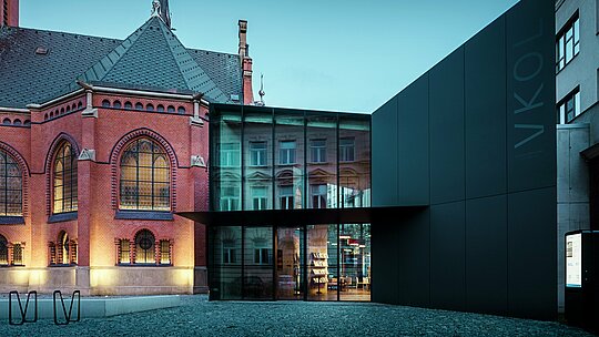 Slika prikazuje impresivno združitev zgodovinske in moderne arhitekture v Olomucu. Izrazita rdeča cerkev v gotskem slogu, vključno z umetelno izdelanimi okni in visokim stolpom, je dopolnjena z moderno novo stavbo z minimalistično črno fasado iz PREFABOND aluminijastih kompozitnih plošč. Prosojne steklene površine nove stavbe odražajo zgodovinsko okolico in ponujajo fascinanten kontrast k trdni strukturi cerkve. Natančna obdelava sodobnih materialov, združena z harmonično povezavo starega in novega, daje stavbi sodobno eleganco, hkrati pa spoštuje zgodovino okolice. Ta kombinacija inovativnih in trajnih materialov, kot je PREFABOND, zagotavlja tako estetsko lepoto kot funkcionalnost.