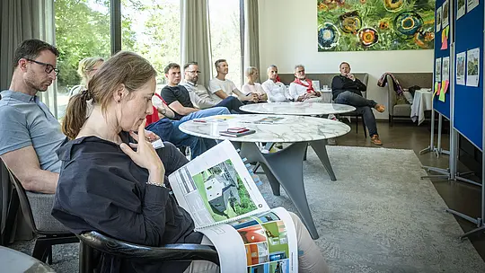 PREFARENZEN 2027: strokovna razprava v svetli seminarski dvorani. Paulina Frey (vodja marketinga PREFA Švica) v rokah drži veliko arhitekturno knjigo s projekti iz PREFA aluminija, medtem ko ji ostali člani žirije pozorno prisluhnejo. Velika okna prepuščajo dnevno svetlobo in odpirajo pogled na zeleno okolico. Prizor izraža zbrano analizo, ustvarjalno razpravo ter vrednotenje oblikovanja in funkcionalnosti. PREFA aluminijasti izdelki simbolizirajo trajne, estetsko dovršene strehe in fasade, ki v arhitekturnih projektih spodbujajo inovativnost, kakovost in navdih.