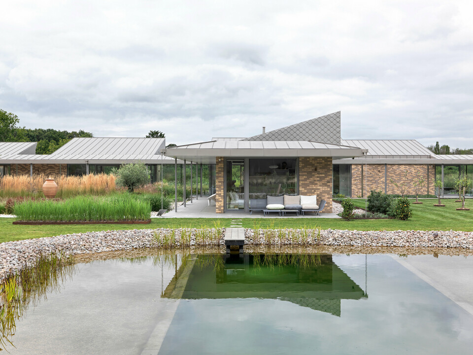 Symmetrische Gesamtansicht des Wohnhauses in West Sussex mit PREFALZ Aluminiumdach in P.10 Bronze und Fassadenakzent mit PREFA Wandrauten 20 × 20. Im Vordergrund ein gestalteter Naturpool mit Holzsteg, umgeben von einer naturnahen Gartenlandschaft. Die klare Architektur mit großflächigen Verglasungen und langlebigen PREFA Materialien schafft ein modernes, witterungsbeständiges und ästhetisch hochwertiges Gebäudeensemble.