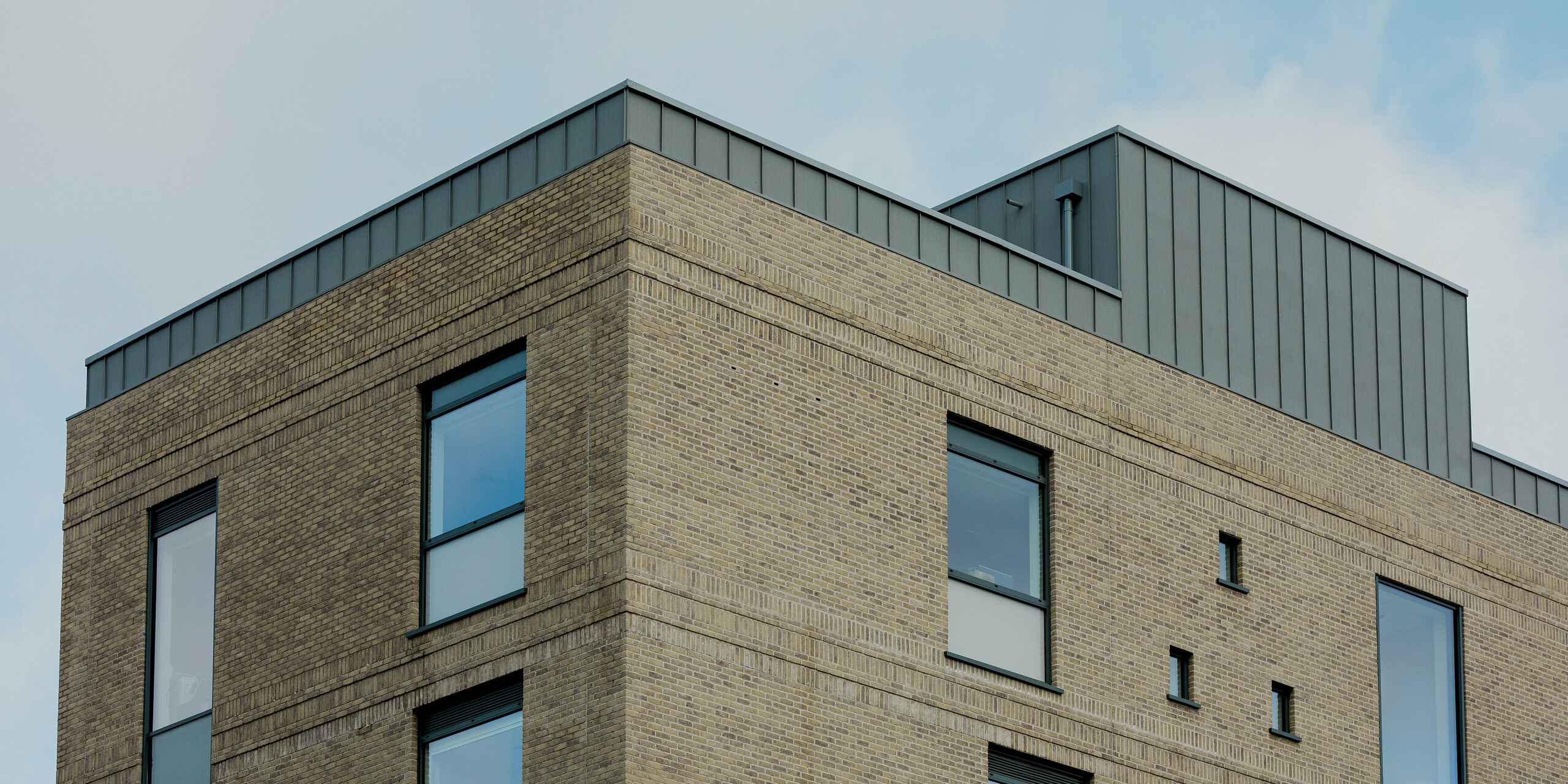 Die klare Gebäudeecke des Northern Regional College in Ballymena tritt mit hellem Klinkermauerwerk und präzisen Fensteröffnungen kraftvoll hervor. Entlang der Dachkante und am zurückgesetzten Aufbau bildet PREFALZ Aluminium in Patinagrau einen markanten, vertikal gegliederten Abschluss. Die Stehfalzbahnen setzen einen ruhigen metallischen Akzent über dem Mauerwerk und betonen die geometrische Architektur. PREFA Aluminium überzeugt durch Langlebigkeit, Witterungsbeständigkeit und eine zeitlose, hochwertige Lösung für moderne Bildungsbauten.