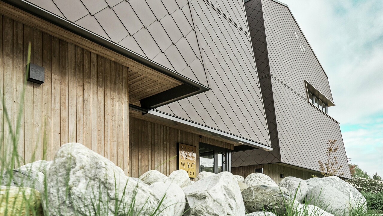 Stranski pogled na hotel, fasada pritličja je vertikalno obložena s temno obarvanim lesom. Nad tem je izstopajoči del fasade, kjer je fasada obložena z aluminijastimi ploščami v orehovo rjavi barvi.