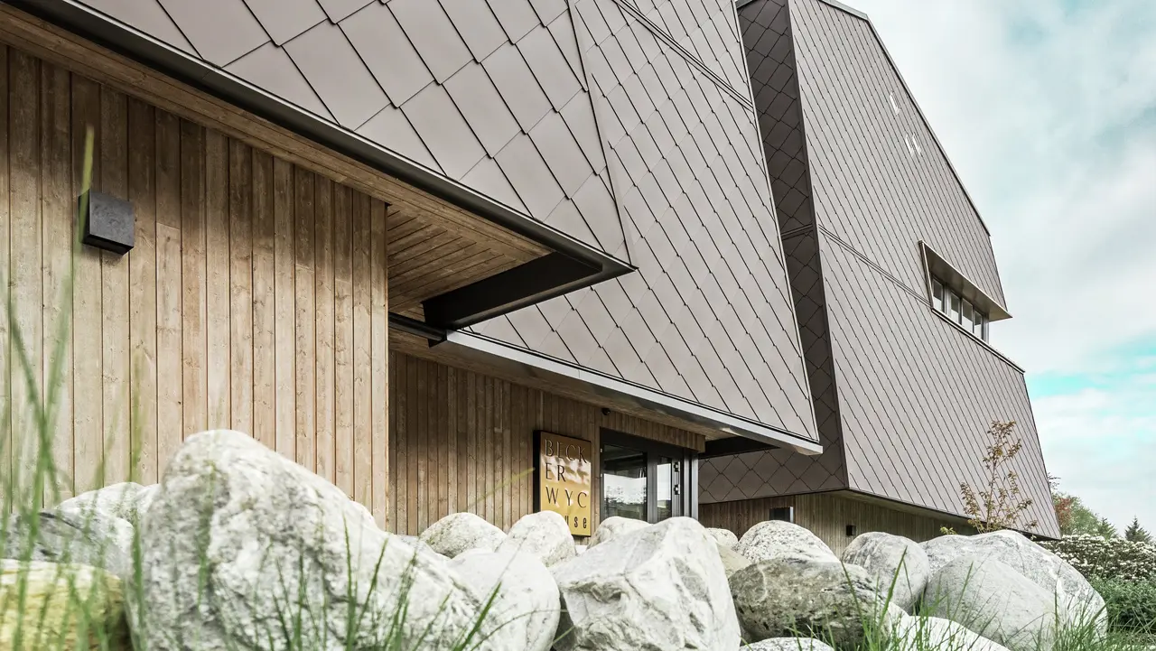 Stranski pogled na hotel, fasada pritličja je vertikalno obložena s temno obarvanim lesom. Nad tem je izstopajoči del fasade, kjer je fasada obložena z aluminijastimi ploščami v orehovo rjavi barvi.