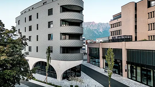Pogled na Bernerhaus v Buchsu z značilno trikotno obliko in nežno zaobljenimi balkoni pred alpskim gorskim ozadjem. Neprekinjena fasada iz PREFA fasadnih skodel v barvi P.10 svetlo siva obdaja stavbo kot luskasta aluminijasta koža in ustvarja živahno igro svetlobe in sence. Nad masivnim betonskim podstavkom z elegantnimi arkadami deluje lahka, vremensko odporna PREFA aluminijasta fasada še posebej natančna, trajna in enostavna za vzdrževanje – arhitekturni magnet za poglede v urbanem okolju. 