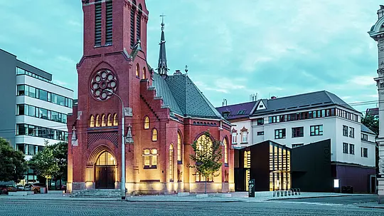  Slika prikazuje harmonično združitev zgodovinske in moderne arhitekture v Olomucu. Impresivna Rdeča cerkev z značilnim koničastim stolpom in gotskimi okni tvori zgodovinsko središče kompleksa. Neposredno ob njej se dviga moderna nova stavba, katere fasada iz matiranih PREFABOND aluminijastih kompozitnih plošč in stekla tvori jasen, eleganten kontrast s tradicionalno arhitekturo. Velike steklene fasade omogočajo vpogled v novo stavbo, ki je na inovativen način povezana s cerkvijo. Sodobna stavba s svojo natančno obliko in jasnimi linijami prispeva k sodobni atmosferi, ne da bi pri tem posegala v zgodovinski značaj cerkve. Ta uspešna simbioza kaže, kako visokokakovostni materiali, kot je PREFABOND, ponujajo popolno podlago za združevanje tradicije in modernosti.