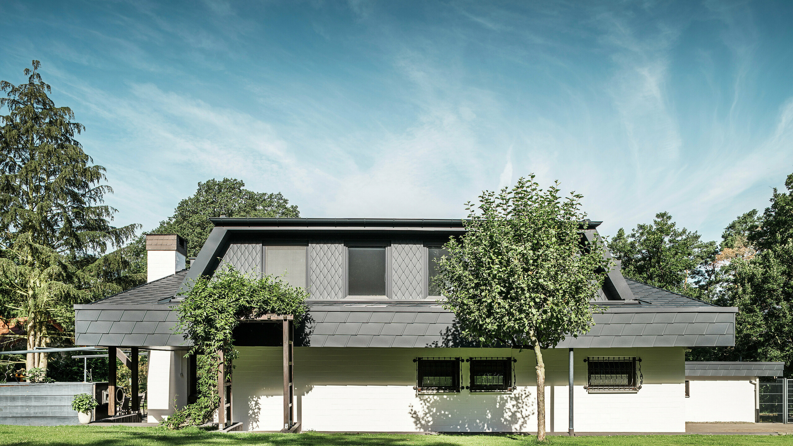Sprednji pogled na vrtno stran enodružinske hiše v Mechernichu z velikim trapezastim strešnim izbočkom, pokritim z majhnimi strešnimi rombi PREFA P.10 antracitne barve. Streha je pokrita s strešnimi skodlami PREFA DS.19 v isti barvi. Fasada spodnjega nadstropja je pobarvana v belo barvo, zelena pergola pa dopolnjuje celotno podobo vrtnega prostora. Drevesa in grmičevje okvirjajo pogled in poskrbijo za harmonično vključitev v naravo.