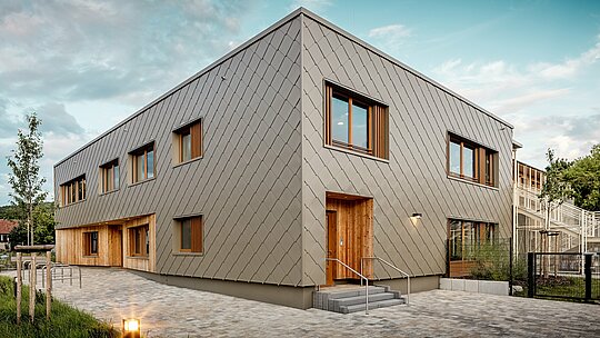 Sodobni vrtec v mestu Alzenau navdušuje s svojo jasno arhitekturo in visokokakovostno PREFA aluminijasto fasado v bronasti barvi. Fasada iz fasadnih rombov 44 × 44 daje stavbi eleganten, strukturiran videz in vizualno povezuje funkcionalne prostore v harmonično celoto. Lesene obloge na vhodih ustvarjajo topel, naraven kontrast z vzdržljivim aluminijem. Kombinacija masivne konstrukcije, lesenih elementov in obešene prezračevane fasade je primer trajnostnega gradbeništva. PREFA ovoj stavbe, ki ne zahteva veliko vzdrževanja, prepriča ne le s svojo reciklabilnostjo, ampak tudi s svojo odpornostjo proti vremenskim vplivom – idealno za prihodnostno usmerjene izobraževalne stavbe z visokimi arhitekturnimi zahtevami.