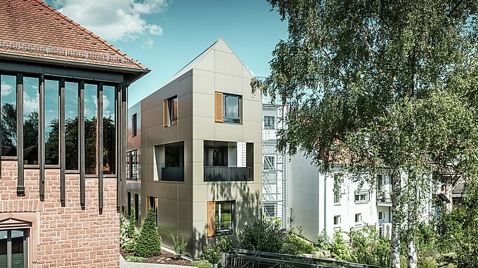 Slika prikazuje moderno mestno hišo v Alzenauu, katere fasada je izdelana iz aluminijastih kompozitnih plošč PREFA v bronasti barvi. Ostrorobi del strehe in jasno strukturirana okna z lesenimi žaluzijami dajejo stavbi elegantno in sodobno estetiko. V ospredju je vidna zelena površina s tlakovano potjo in drevesi, ki harmonično dopolnjuje urbano obliko stavbe.