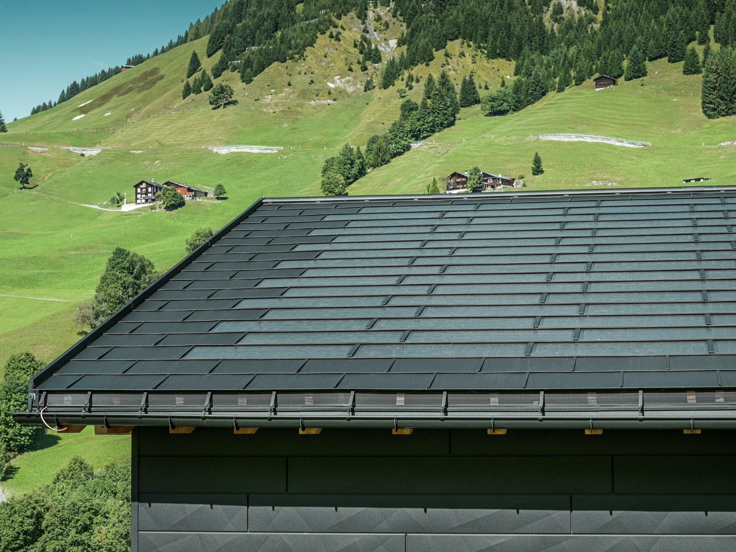 Približek strehe moderne enodružinske hiše v alpskem okolju v Ascharini v Švici. Streha je pokrita z inovativno solarno strešno ploščo PREFA in tradicionalno strešno ploščo R.16, obe v barvi P.10 črna. Za zanesljivo odvodnjavanje strehe skrbijo strešni žleb PREFA in iztočna cev PREFA. Na spodnjem robu slike so vidni prepoznavni fasadni paneli PREFA Siding.X. Jasne linije aluminijastih izdelkov poudarjajo sodoben dizajn sredi slikovite gorske pokrajine.