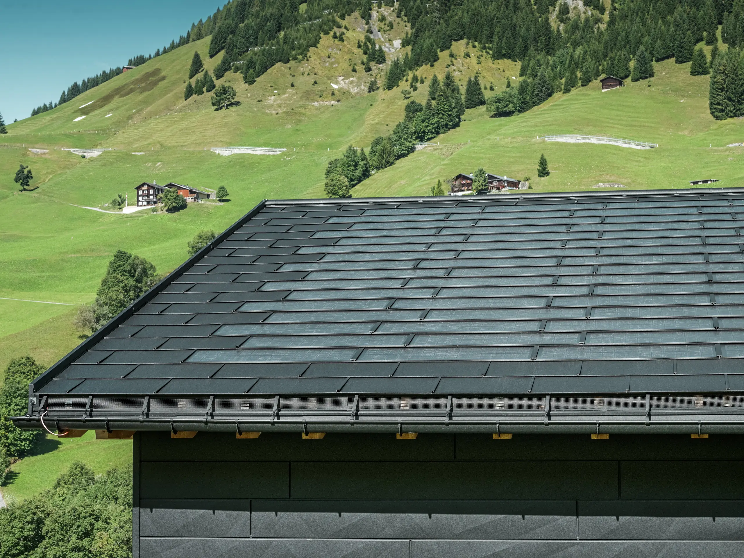 Približek strehe moderne enodružinske hiše v alpskem okolju v Ascharini v Švici. Streha je pokrita z inovativno solarno strešno ploščo PREFA in tradicionalno strešno ploščo R.16, obe v barvi P.10 črna. Za zanesljivo odvodnjavanje strehe skrbijo strešni žleb PREFA in iztočna cev PREFA. Na spodnjem robu slike so vidni prepoznavni fasadni paneli PREFA Siding.X. Jasne linije aluminijastih izdelkov poudarjajo sodoben dizajn sredi slikovite gorske pokrajine.
