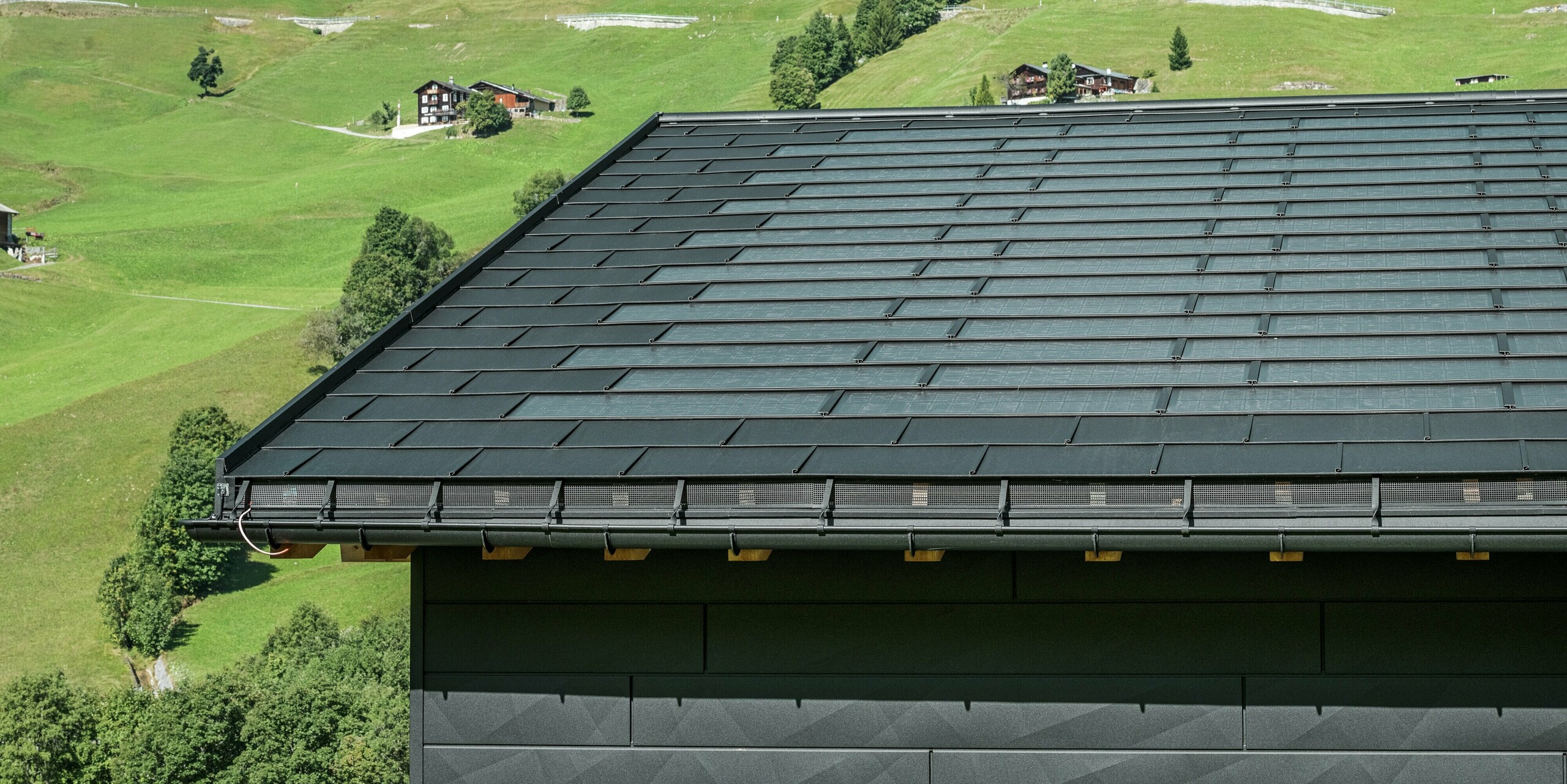 Približek strehe moderne enodružinske hiše v alpskem okolju v Ascharini v Švici. Streha je pokrita z inovativno solarno strešno ploščo PREFA in tradicionalno strešno ploščo R.16, obe v barvi P.10 črna. Za zanesljivo odvodnjavanje strehe skrbijo strešni žleb PREFA in iztočna cev PREFA. Na spodnjem robu slike so vidni prepoznavni fasadni paneli PREFA Siding.X. Jasne linije aluminijastih izdelkov poudarjajo sodoben dizajn sredi slikovite gorske pokrajine.