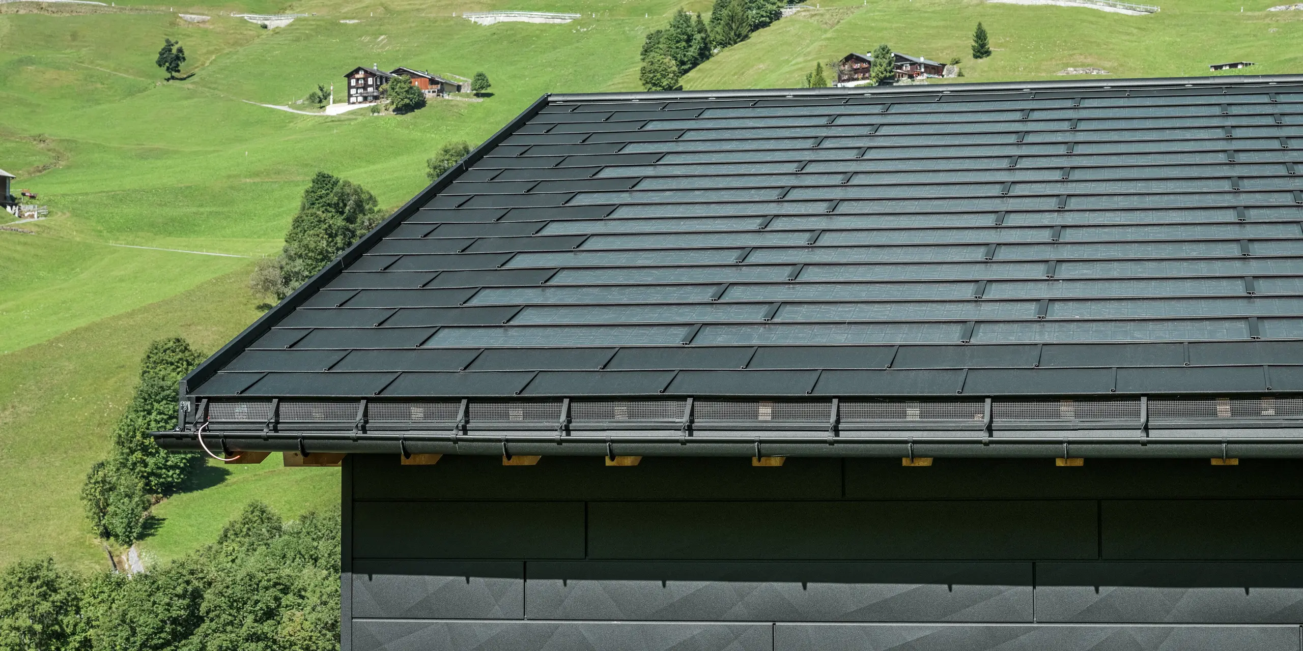 Približek strehe moderne enodružinske hiše v alpskem okolju v Ascharini v Švici. Streha je pokrita z inovativno solarno strešno ploščo PREFA in tradicionalno strešno ploščo R.16, obe v barvi P.10 črna. Za zanesljivo odvodnjavanje strehe skrbijo strešni žleb PREFA in iztočna cev PREFA. Na spodnjem robu slike so vidni prepoznavni fasadni paneli PREFA Siding.X. Jasne linije aluminijastih izdelkov poudarjajo sodoben dizajn sredi slikovite gorske pokrajine.