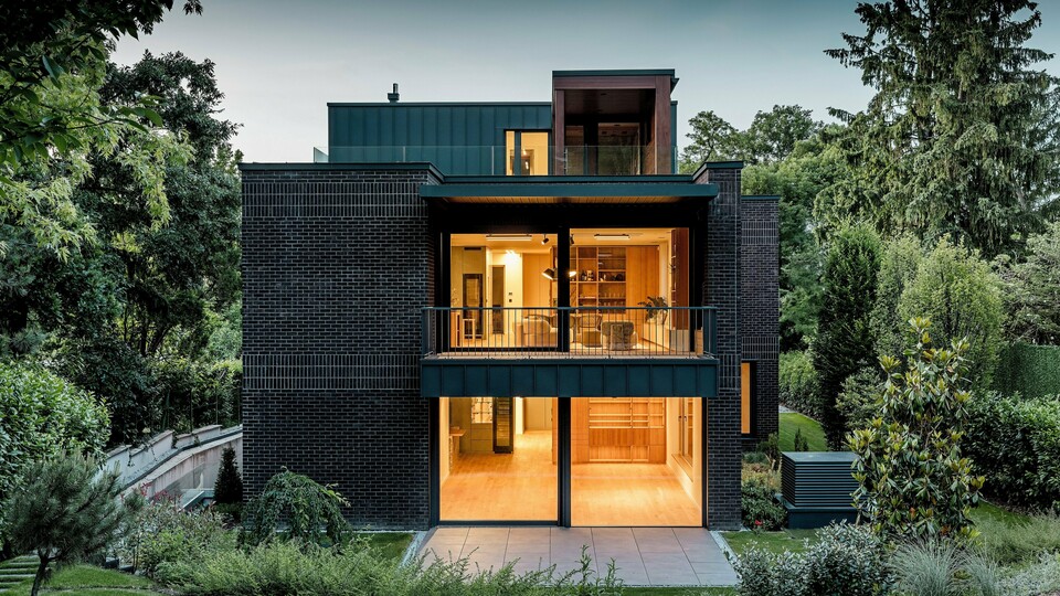 Fotografija prikazuje moderno zadnjo stran večgeneracijske vile v Budimpešti v mraku. Prostorne steklene fasade odpirajo pogled na toplo osvetljene notranje prostore, medtem ko kombinacija temno rdeče opečne fasade in aluminija PREFALZ v barvi P.10 antracit poudarja jasne arhitekturne linije. Natančno izvedeni kotni stoječi zgibi poudarjajo visokokakovostno izdelavo in dolgo življenjsko dobo PREFA aluminijastih izdelkov. Obdana z bujno zeleno vegetacijo se stavba harmonično vklaplja v krajino vrta in izžareva eleganten, sodoben karakter. 