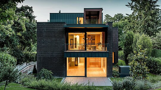 Fotografija prikazuje moderno zadnjo stran večgeneracijske vile v Budimpešti v mraku. Prostorne steklene fasade odpirajo pogled na toplo osvetljene notranje prostore, medtem ko kombinacija temno rdeče opečne fasade in aluminija PREFALZ v barvi P.10 antracit poudarja jasne arhitekturne linije. Natančno izvedeni kotni stoječi zgibi poudarjajo visokokakovostno izdelavo in dolgo življenjsko dobo PREFA aluminijastih izdelkov. Obdana z bujno zeleno vegetacijo se stavba harmonično vklaplja v krajino vrta in izžareva eleganten, sodoben karakter. 