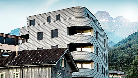 Moderna, trikotna stanovanjsko-poslovna stavba v Buchsu z izrazito zaobljeno arhitekturo in fasado v obliki skodle, obloženo s PREFA fasadnimi skodlami v barvi P.10 svetlo siva. Enakomerno obdajajoča aluminijasta fasada ustvarja živahen videz, ki spominja na plazilce, in se kot eleganten poudarek izpostavlja pred alpskim gorskim ozadjem. Zahvaljujoč lahkemu, vremensko odporenemu in trajnemu PREFA aluminiju so natančno izvedeni tudi ozki radiji in zaobljenosti – za homogeno, vzdrževanju prijazno ovojnico stavbe z brezčasno moderno izžarevanostjo.