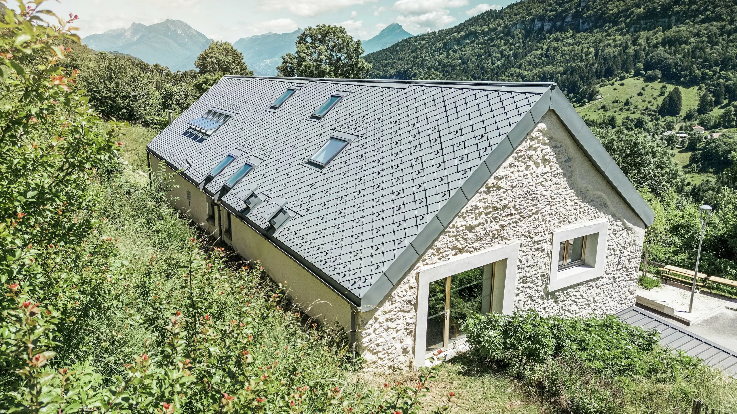 Pogled na streho prenovljenega skednja „La Grange à Gaby“ v Engins v Franciji. Stavba je pokrita s trajnimi strešnimi rombi PREFA 29 × 29, ki združujejo sodobno zaščito s tradicionalno estetiko. Idilična gorska pokrajina poudarja harmonično vpetost v naravo.
