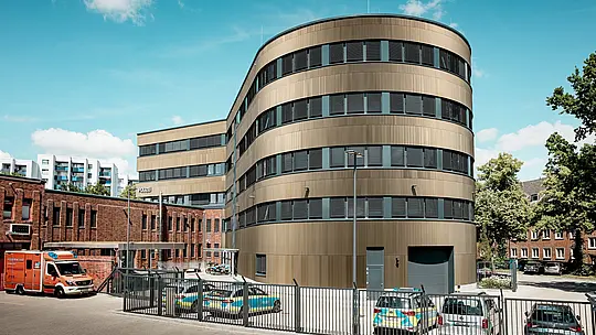 Značilen zunanji videz policijske postaje 43 v Hamburgu: ukrivljena nova stavba s PREFA aluminijasto fasado iz eloksiranih ekstrudiranih zobčastih profilov v barvi srednje bronaste deluje kot močan magnet za poglede v mestni krajini. Fina, navpična struktura ustvarja živahne svetlobne in senčne učinke ter poudarja zaobljeno arhitekturo. V kontrastu s tem stoji zgodovinska opekarna. PREFA aluminij prepriča z visoko odpornostjo proti vremenskim vplivom, zaščito pred korozijo, dolgo življenjsko dobo in minimalnimi stroški vzdrževanja – idealno za trajnostne, funkcionalne in estetske fasadne rešitve.