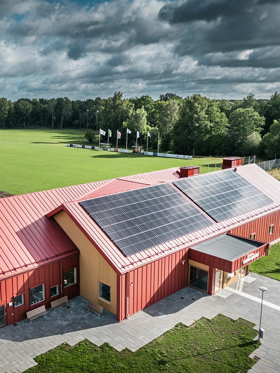 Luftaufnahme der Schulkantine Restaurang Gröna Hjärtat in Broby, Schweden, zeigt das Gebäude mit auffälliger Stehfalzdeckung am Dach und der Fassade aus PREFALZ in Oxydrot. Auf dem Dach ist eine großflächige Photovoltaikanlage installiert, die Strom aus erneuerbarer Energie für das Gebäude liefert. Die klare architektonische Linienführung wird durch die kontrastreiche Kombination aus rotem Aluminium und warmem Holz an den Giebelseiten unterstrichen. Im Hintergrund erstreckt sich ein großes Sportfeld, das in die grüne Umgebung eingebettet ist. Diese Aufnahme betont die harmonische Integration des modernen Gebäudes in die natürliche Landschaft.