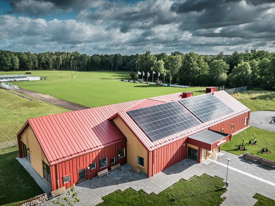 Luftaufnahme der Schulkantine Restaurang Gröna Hjärtat in Broby, Schweden, zeigt das Gebäude mit auffälliger Stehfalzdeckung am Dach und der Fassade aus PREFALZ in Oxydrot. Auf dem Dach ist eine großflächige Photovoltaikanlage installiert, die Strom aus erneuerbarer Energie für das Gebäude liefert. Die klare architektonische Linienführung wird durch die kontrastreiche Kombination aus rotem Aluminium und warmem Holz an den Giebelseiten unterstrichen. Im Hintergrund erstreckt sich ein großes Sportfeld, das in die grüne Umgebung eingebettet ist. Diese Aufnahme betont die harmonische Integration des modernen Gebäudes in die natürliche Landschaft.