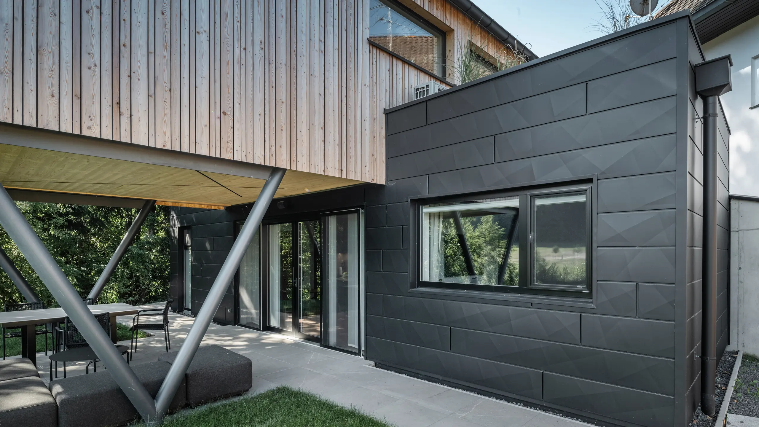 Arhitekturni vrhunec: „Woodstock Chalet 5” v St. Martin am Innkreis, Zgornja Avstrija. Sodobno namestitev za poroke poudarjajo fasadni paneli PREFA Siding.X v antracitni barvi. Aluminijasti elementi tvorijo močan kontrast s svetlimi lesnimi ploščami in kovinskimi nosilnimi konstrukcijami. Temni okenski okvirji se harmonično vključujejo v oblikovanje fasade, kar daje stavbi eleganten videz. Ta slika prikazuje estetsko ravnovesje med naravno teksturo lesa in industrijskim šarmom aluminijaste fasade, ki predstavlja oblikovanje sodobnih stavb.