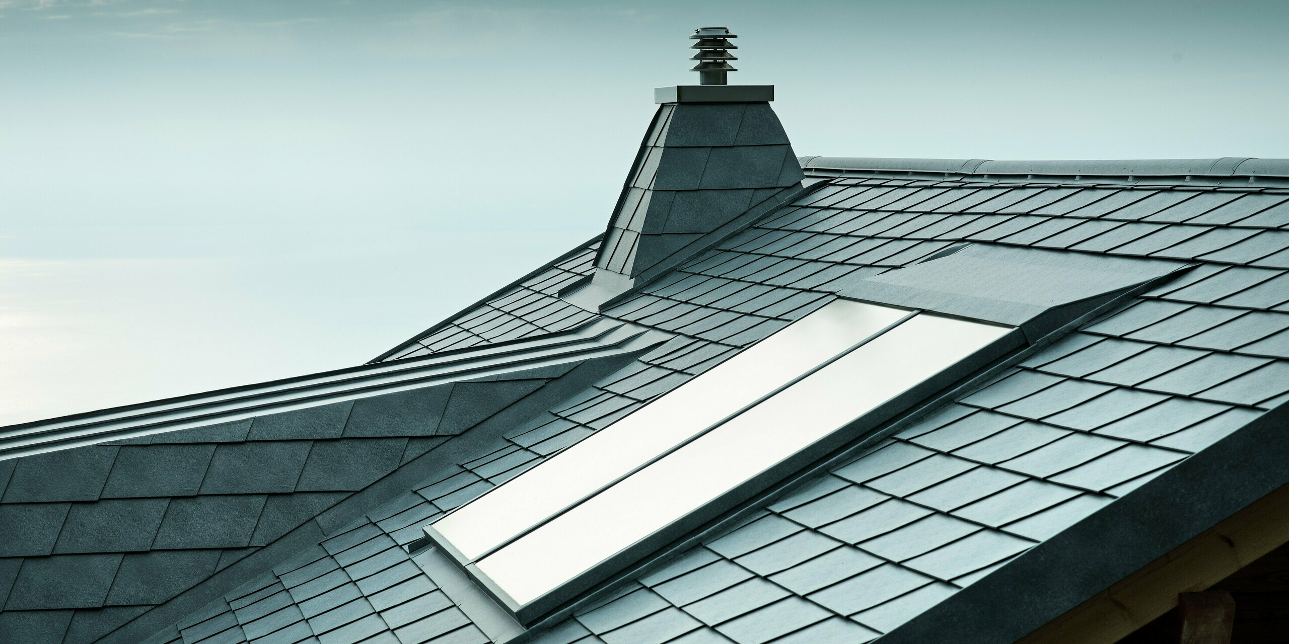 Diese vielseitige Dachansicht zeigt steingraue PREFA-Dachschindeln sowie einen Kamin in Trapezform aus Aluminium. Das langlebige Aluminiumdach ist nicht nur stilvoll, sondern auch äußerst widerstandsfähig. Die exakte Ausführung der Dachgaube und des Dachfensters zeugt von der Detailgenauigkeit der PREFA-Verlegepartner.